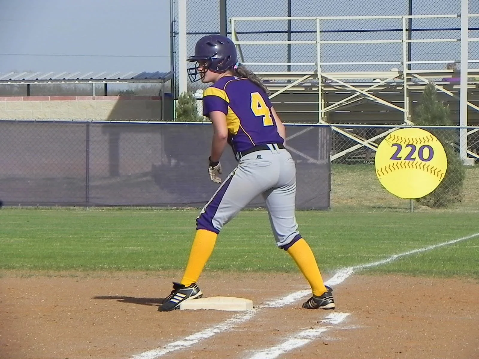 A female softball player wearing a purple and yellow uniform with the number 4, standing on a base during a game, on a softball field.