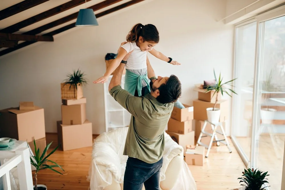 A man lifting a young girl in a living room with moving boxes, plants, and large windows.