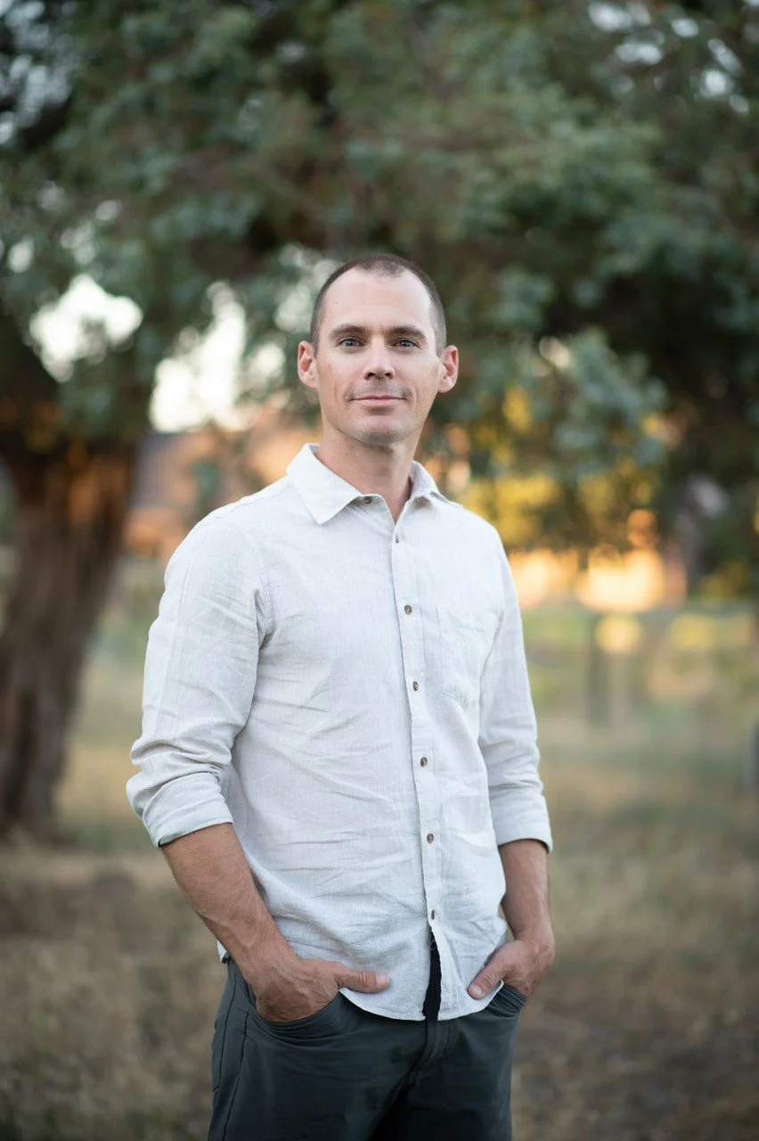 A Josh Garner the founder wearing a white button-up shirt and dark pants stands outdoors with hands in pockets, with trees in the background.