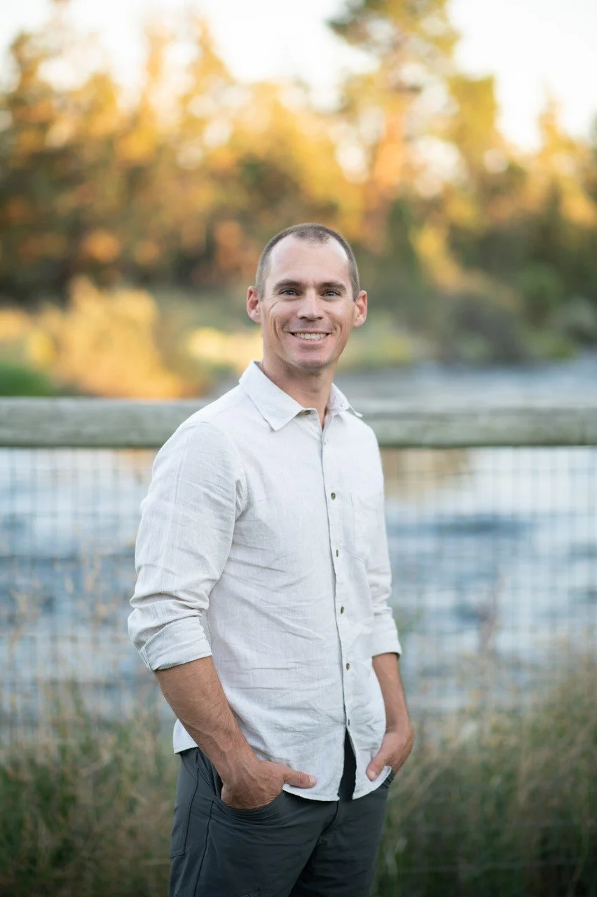 A Josh Garner the founder wearing a white button-up shirt and dark pants stands outdoors with hands in pockets, a wooden fence and trees in the background.
