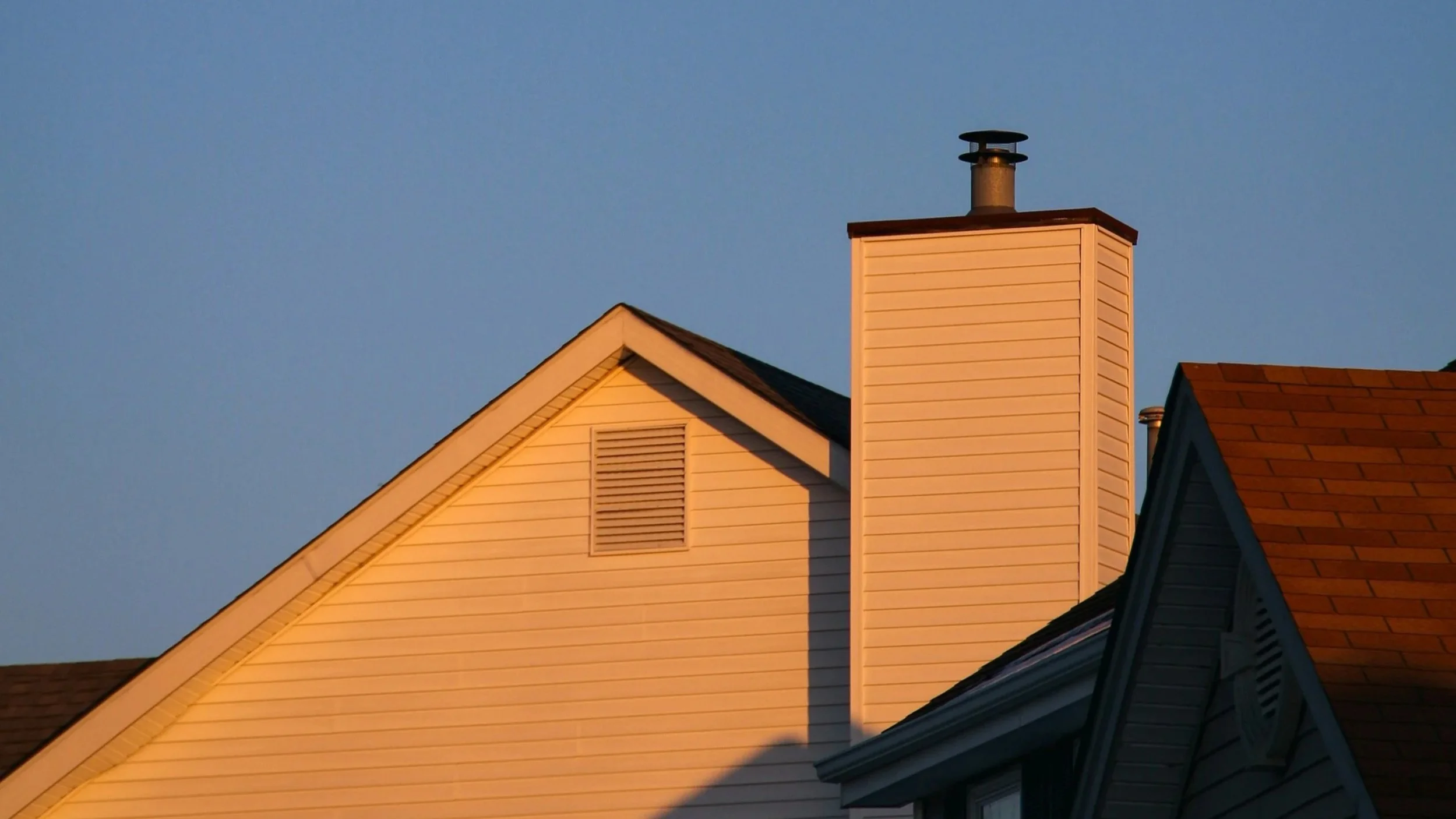 Close-up of house rooftops with beige and brown shingles, chimney, and gable, under a clear blue sky.