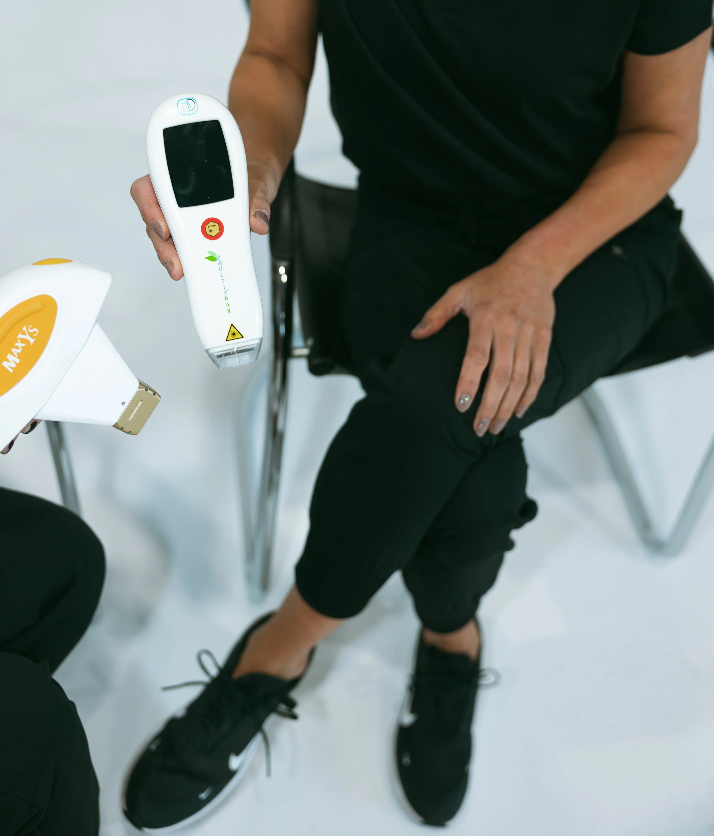 Person sitting with legs crossed, wearing black sneakers and black clothing, holding a white handheld device with a screen and yellow and red labels, on a clean, white floor.