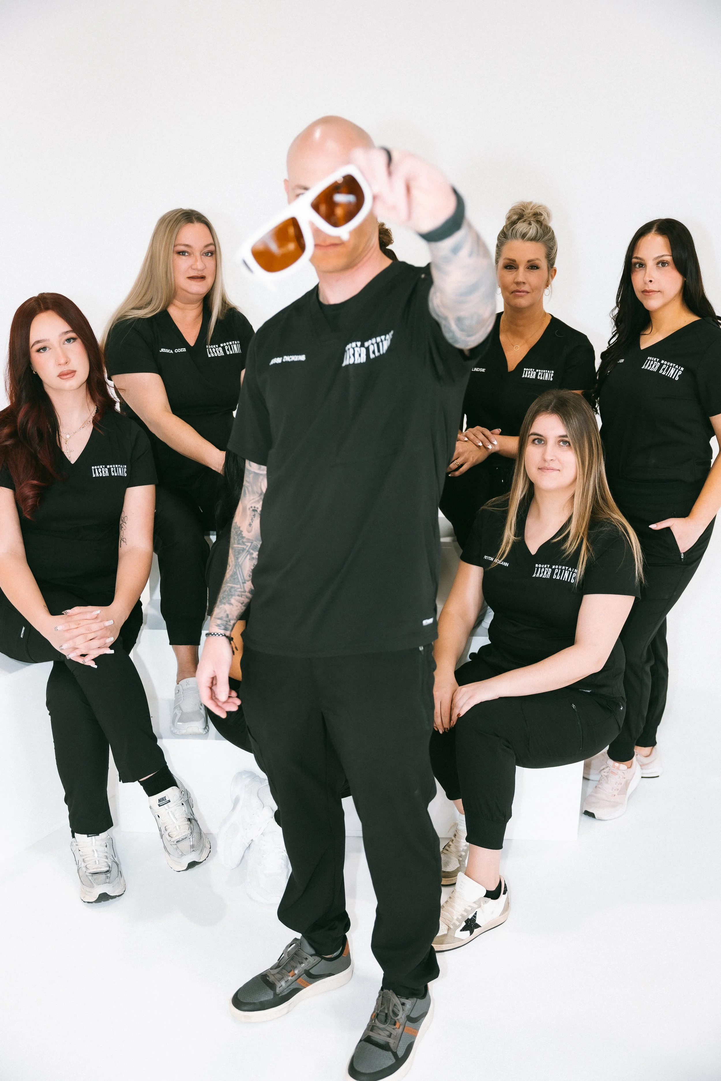 A group of six women and one man posing in a white studio. The man in the center is holding a pair of sunglasses toward the camera. All are wearing black shirts with white text and black pants or leggings.