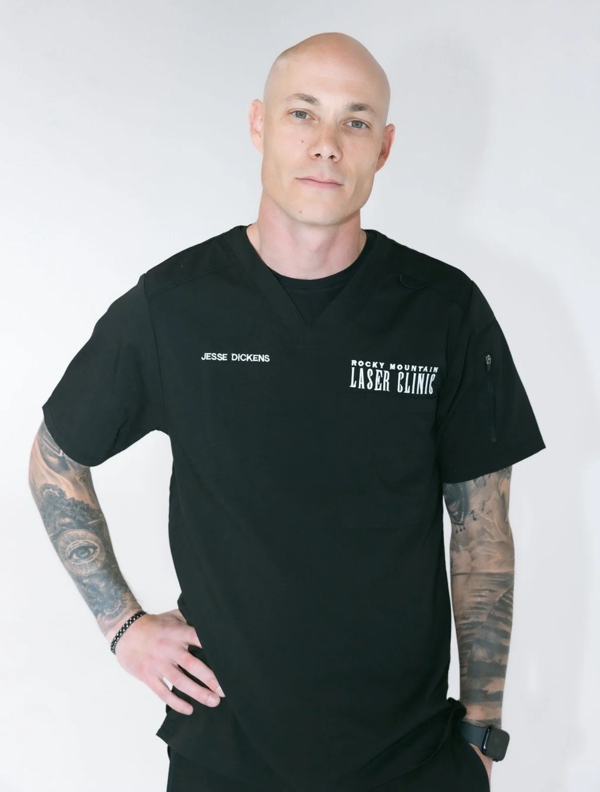 A bald man in black medical scrubs with tattoos on his arms, standing against a white background. His scrubs have text that reads Jesse Dickens and Rocky Mountain Laser Clinic.