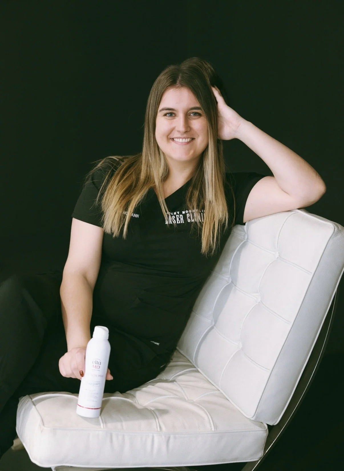 A young woman with long light brown hair, wearing a black t-shirt, sitting on a white cushioned chair against a black background. She is smiling, holding a white bottle of lotion or skincare product in her right hand, and her left hand is resting on her head.