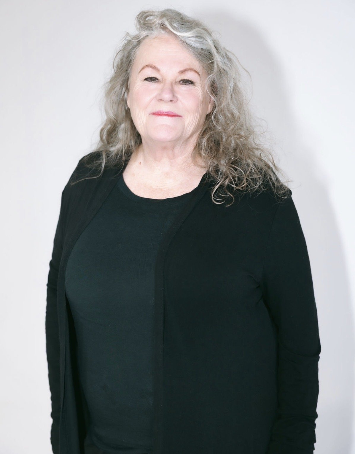 An elderly woman with long, curly gray hair, wearing a black top and black cardigan, standing against a plain white background.