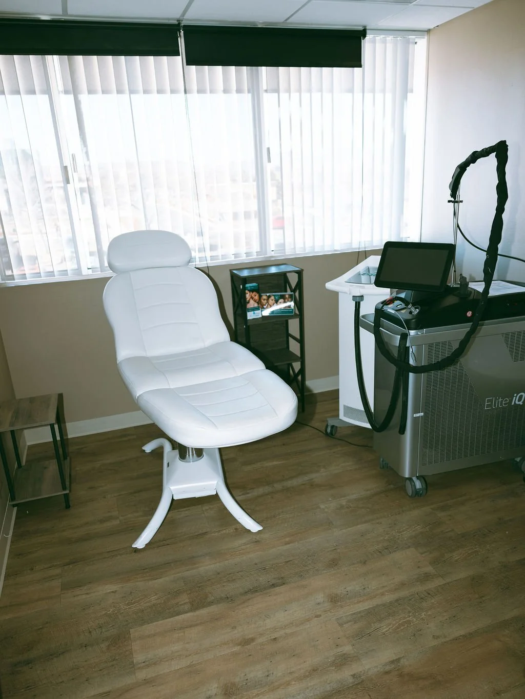 Medical examination room with a white chair, a small black side table with pamphlets, a medical device with a screen, and a window with blinds.