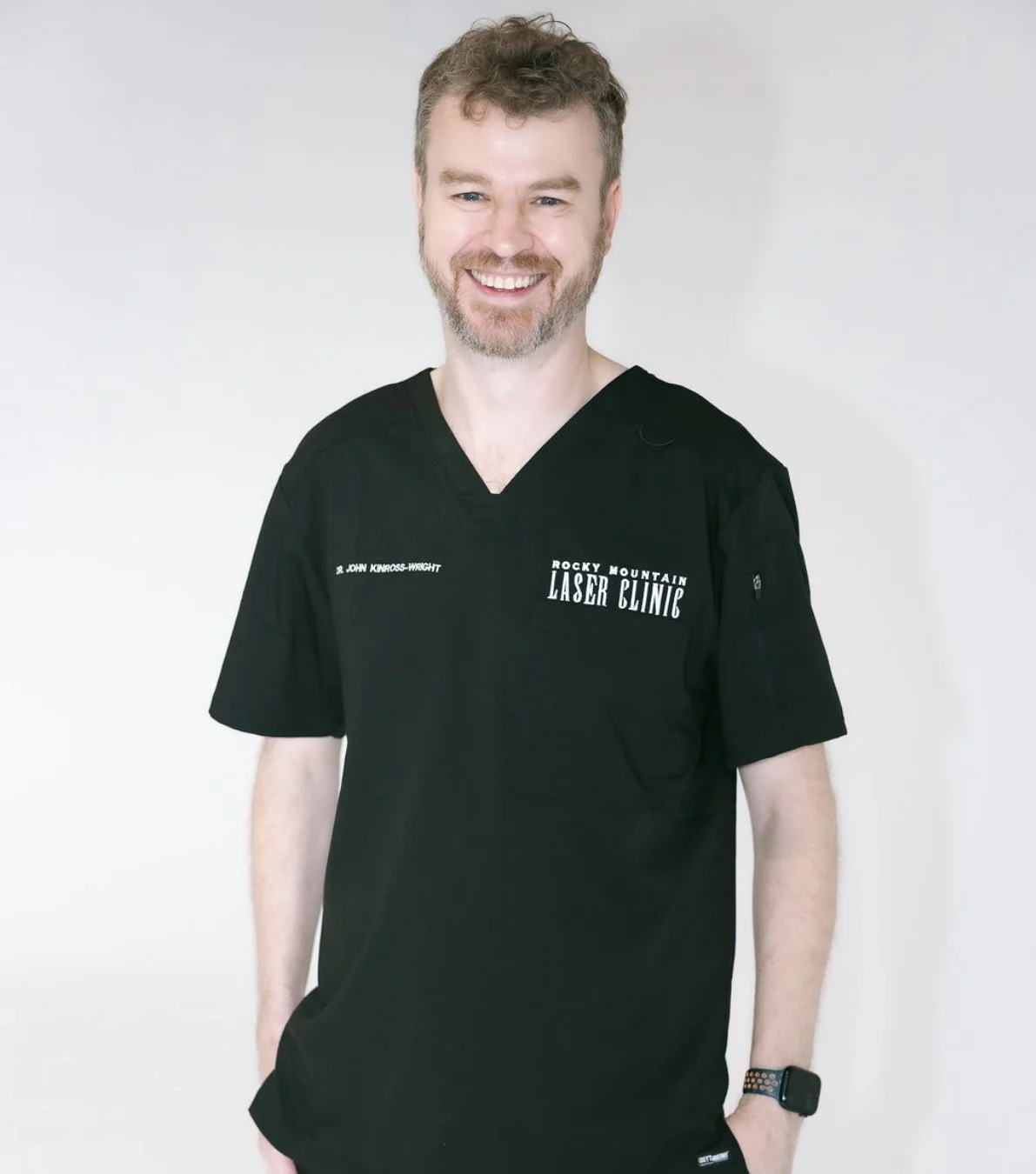 A smiling man wearing black medical scrubs with 'Rocky Mountain Laser Clinic' and 'Dr. John Kinross-Wright' written on it, standing against a plain light background.