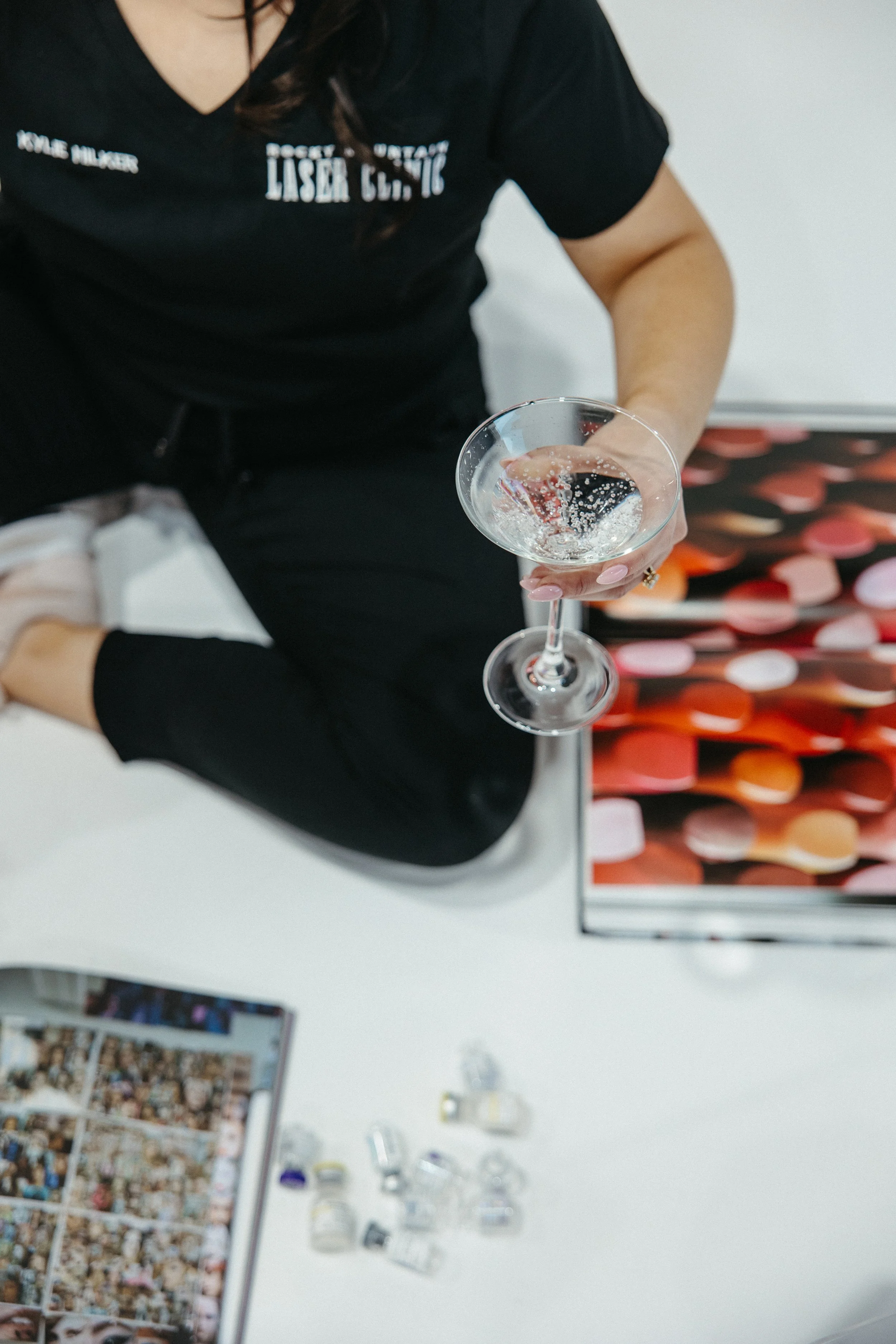 A woman in a black t-shirt with white text is holding a martini glass with a bubbly clear beverage.