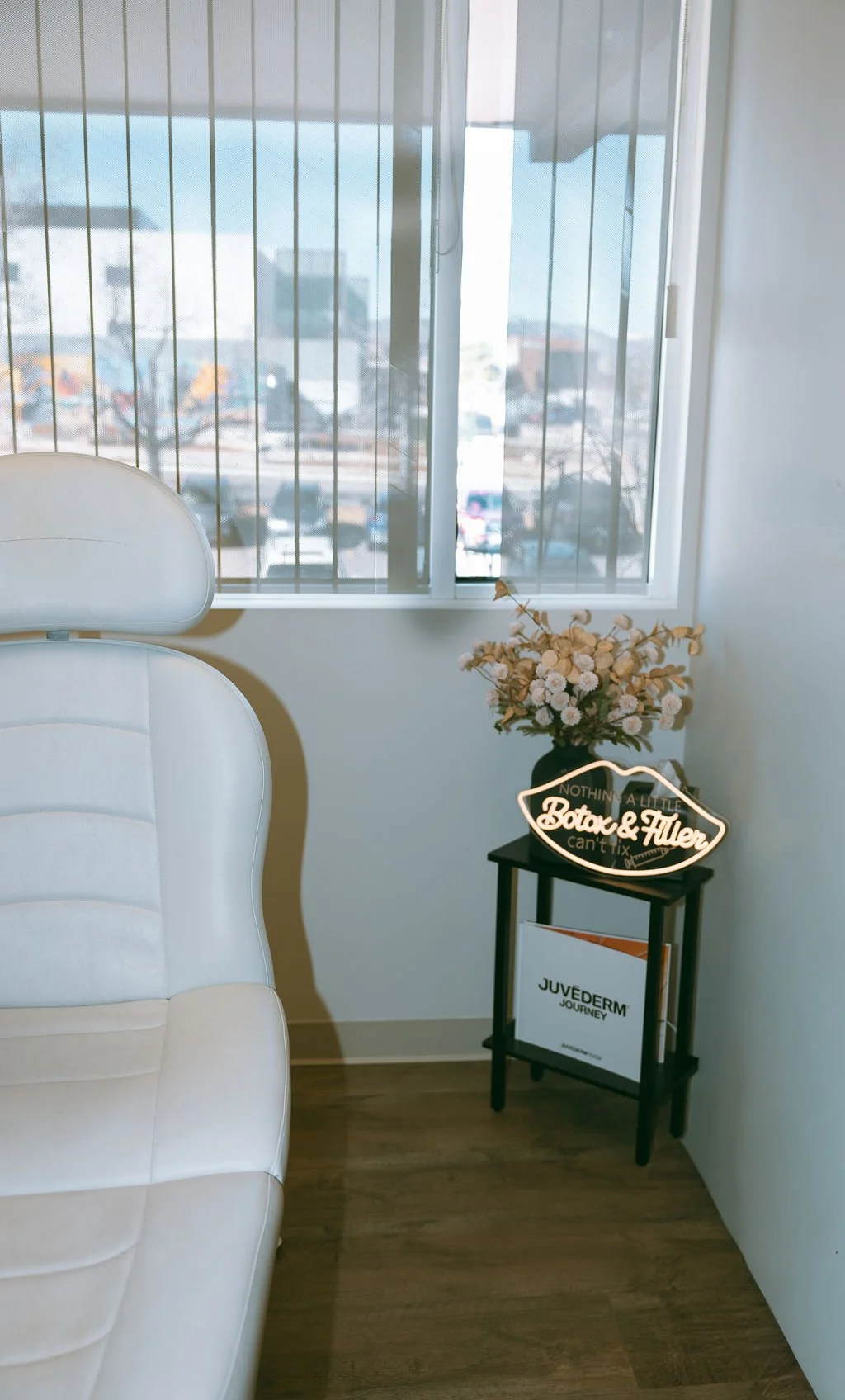 Part of a white chair, a small side table with a flower arrangement, a neon sign, and a window with metal bars overlooking a city street.