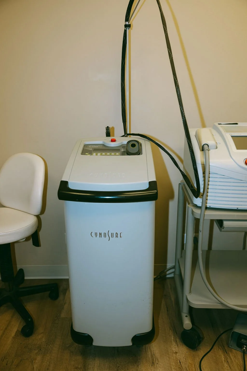 Medical laser device labeled 'Cynosure' beside a white chair and medical equipment on a trolley in a clinical setting.