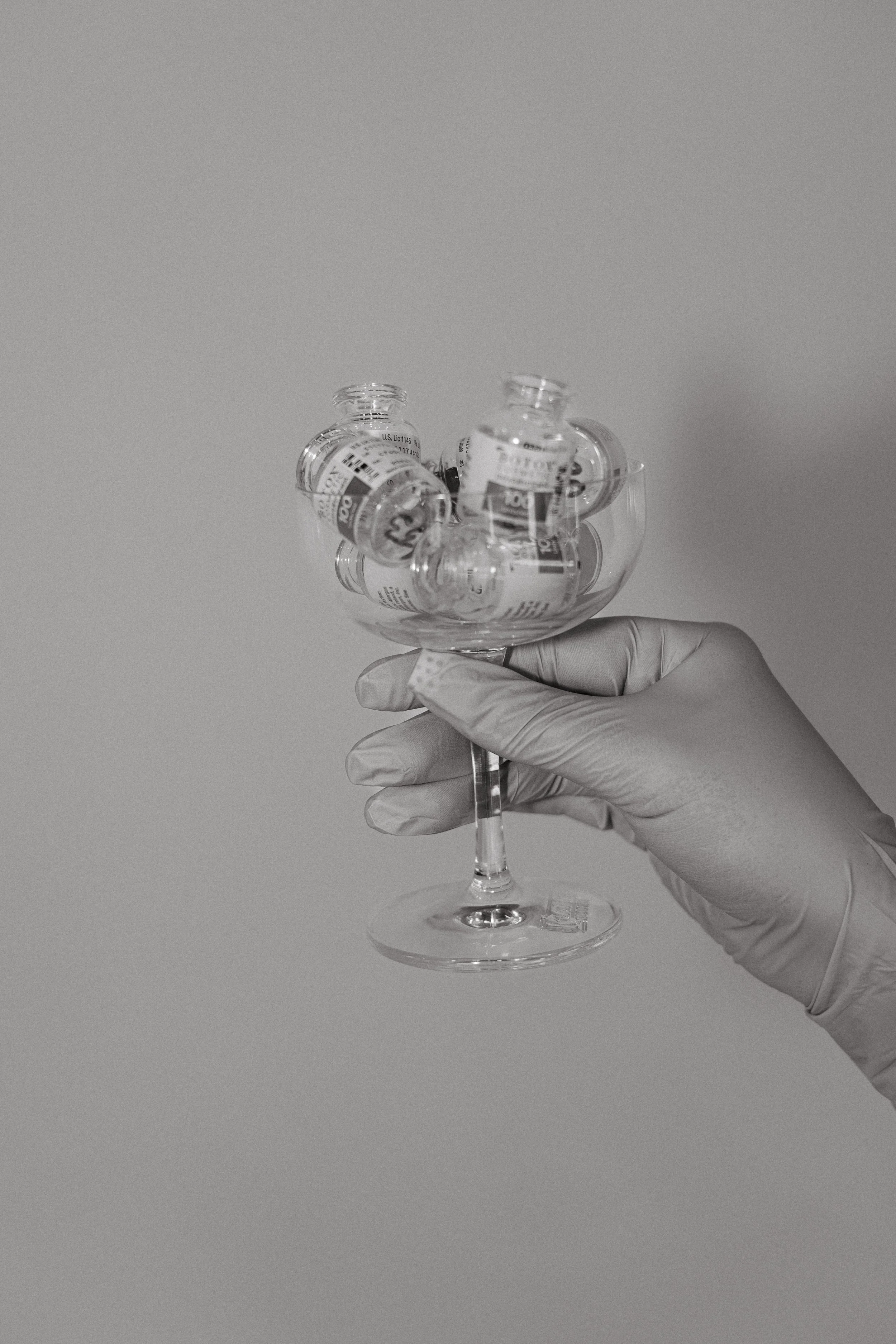 Hand wearing a medical glove holding a glass container filled with small glass vials, against a plain background.