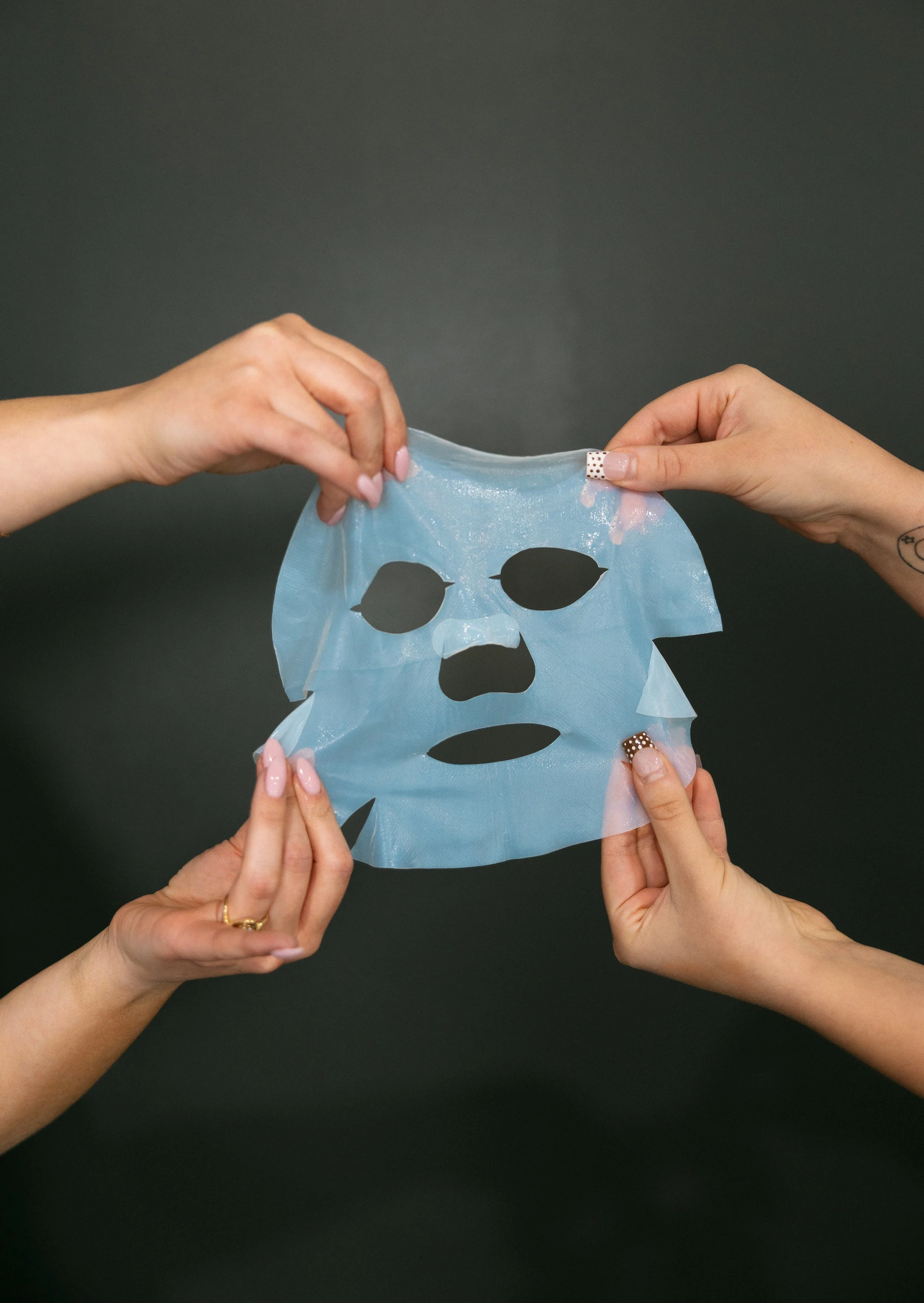 Multiple hands are holding a blue sheet mask with cutouts for eyes, nose, and mouth against a dark background.
