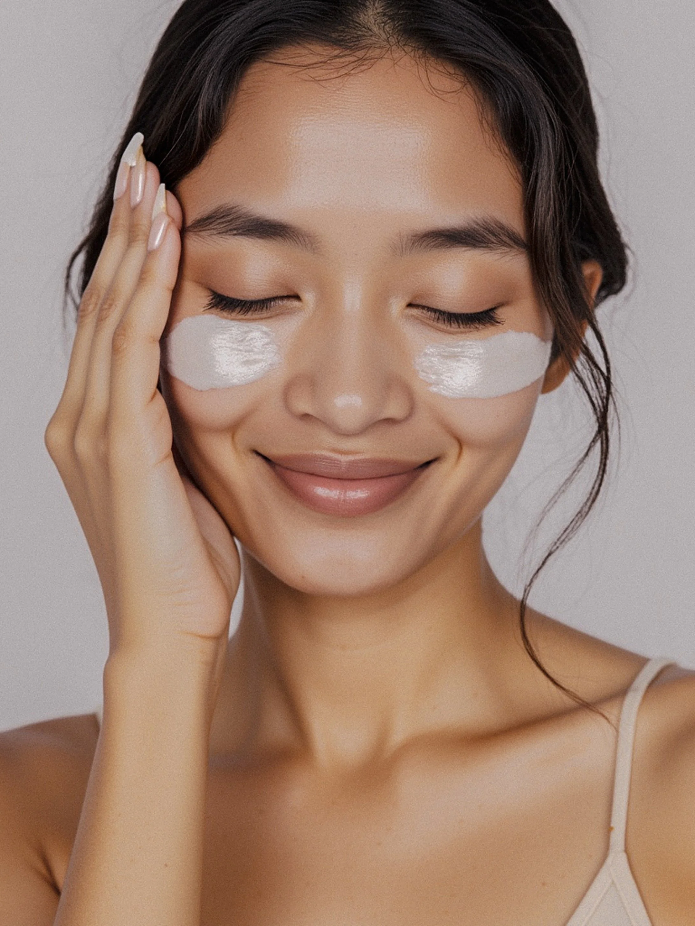A woman with dark hair and light skin applying white facial cream under her eyes, smiling gently with her eyes closed.