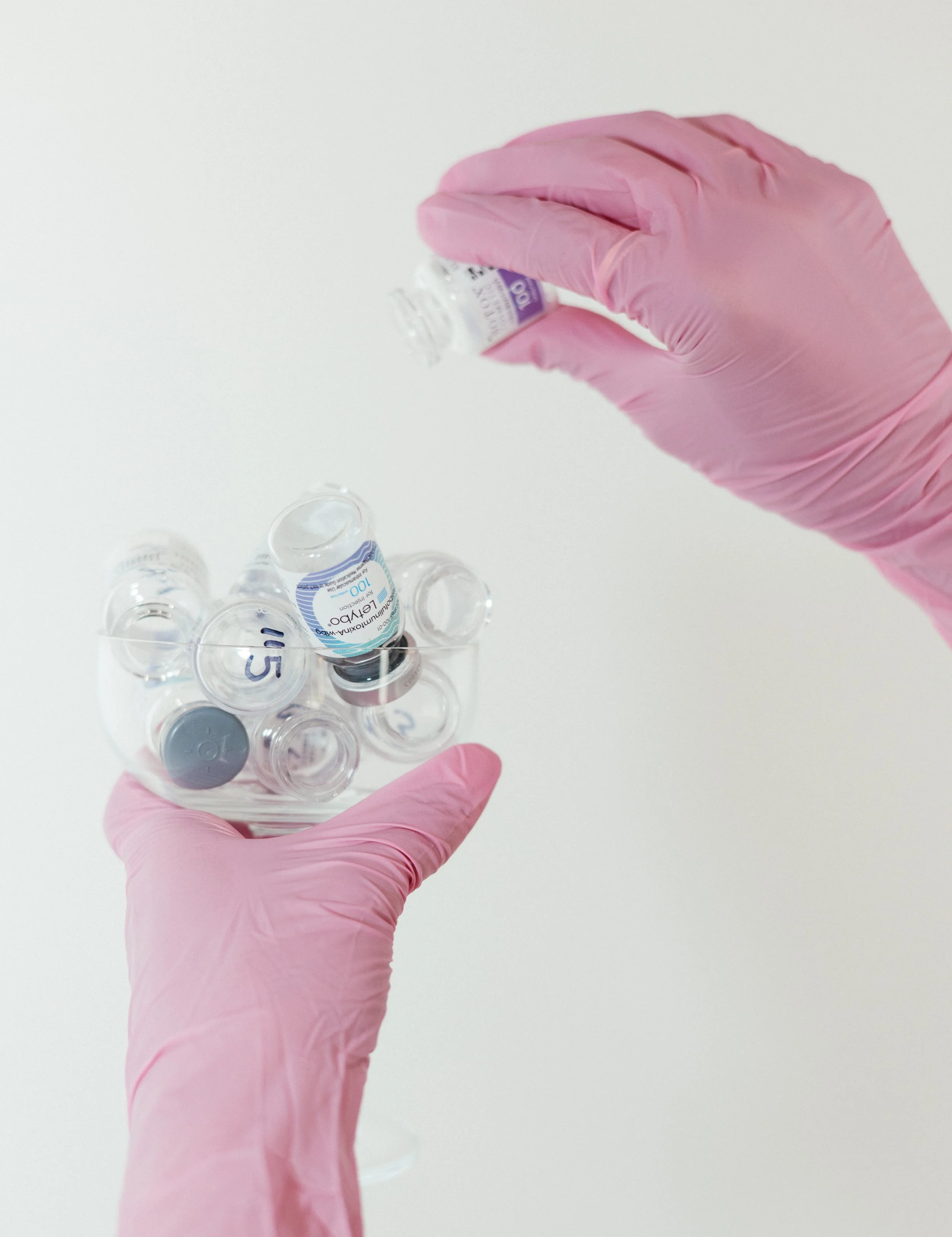 A person wearing pink gloves holding a small vial of COVID-19 vaccine with other vials placed on a white surface.