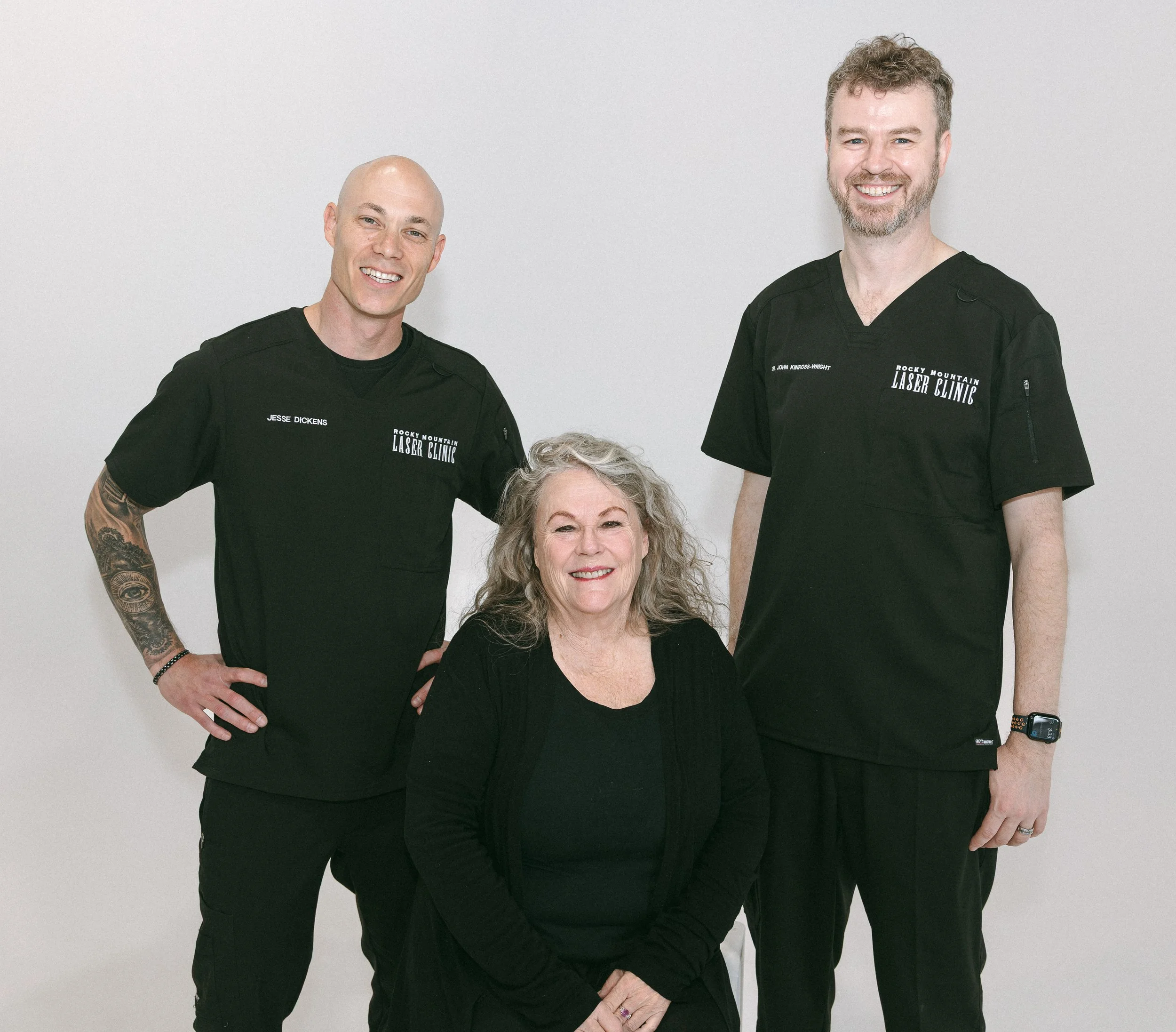 Two smiling male healthcare professionals dressed in black scrubs, standing against a plain white background, with a partially visible older woman in front of them.