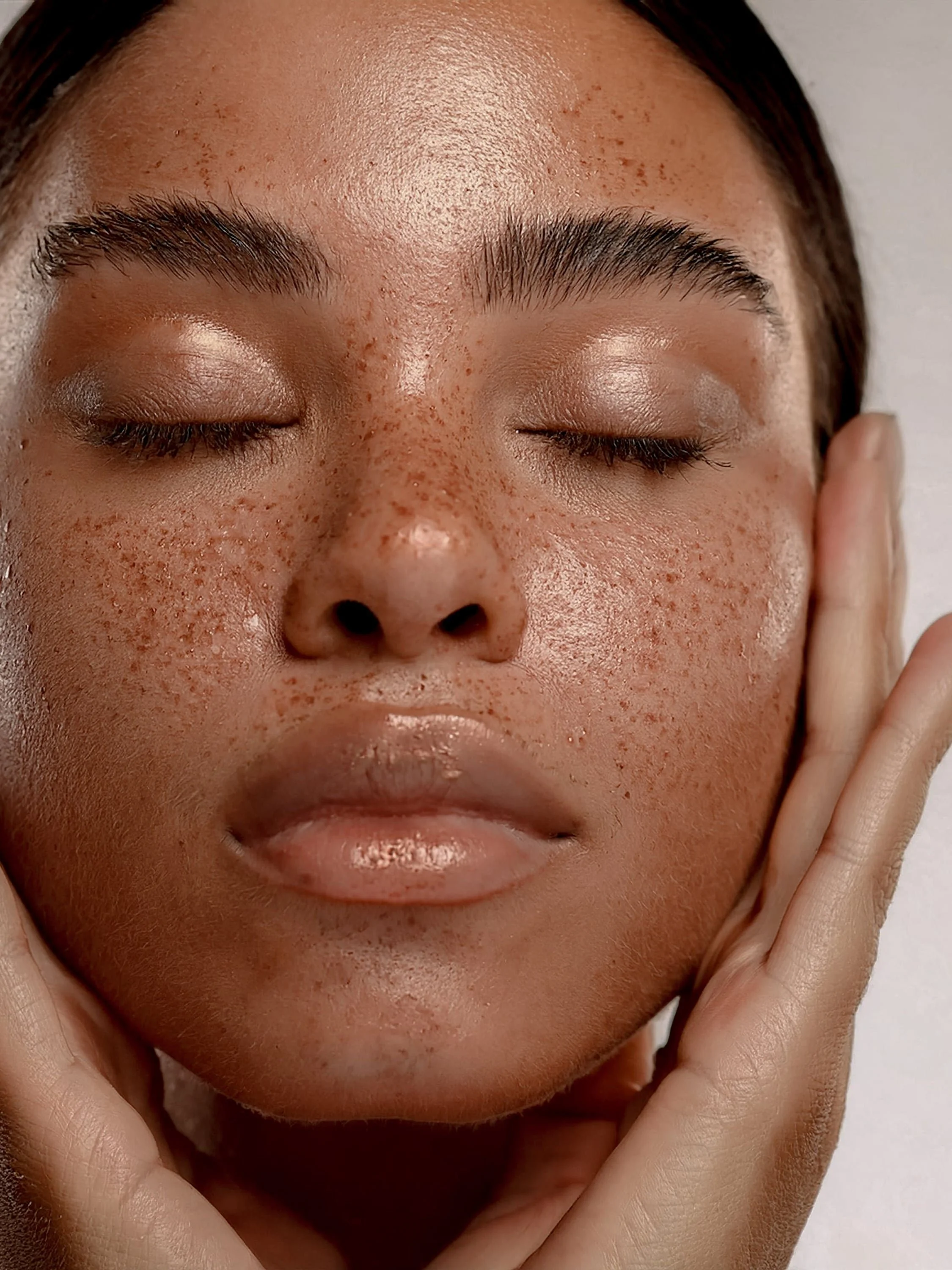 Close-up of a person's face with closed eyes, showing freckles and natural skin texture, gently touching their face with fingers.