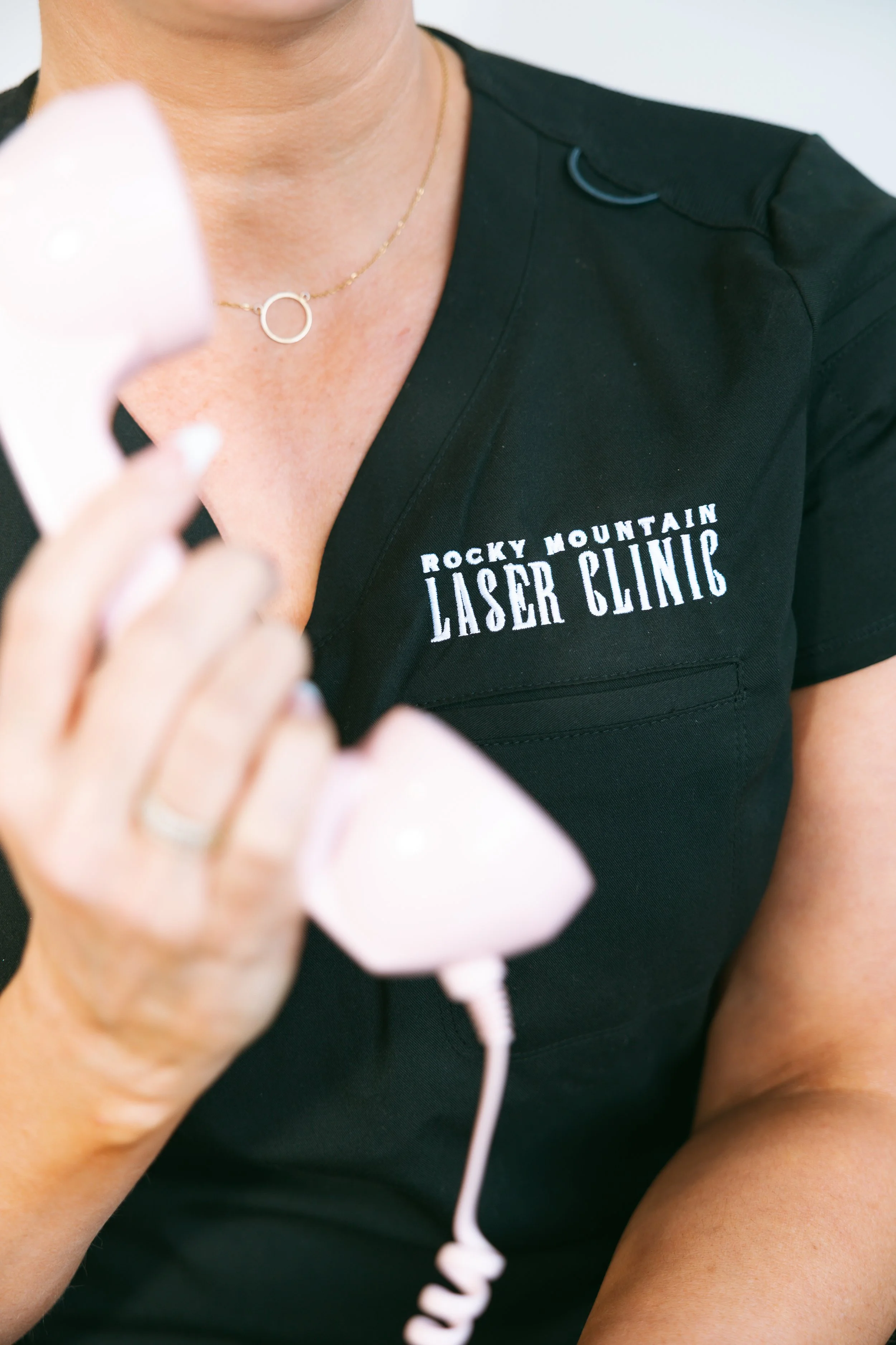 A person wearing a black shirt with 'Rocky Mountain Laser Clinic' embroidered on it, holding a pink ultrasound probe, with a gold necklace featuring a circular pendant.