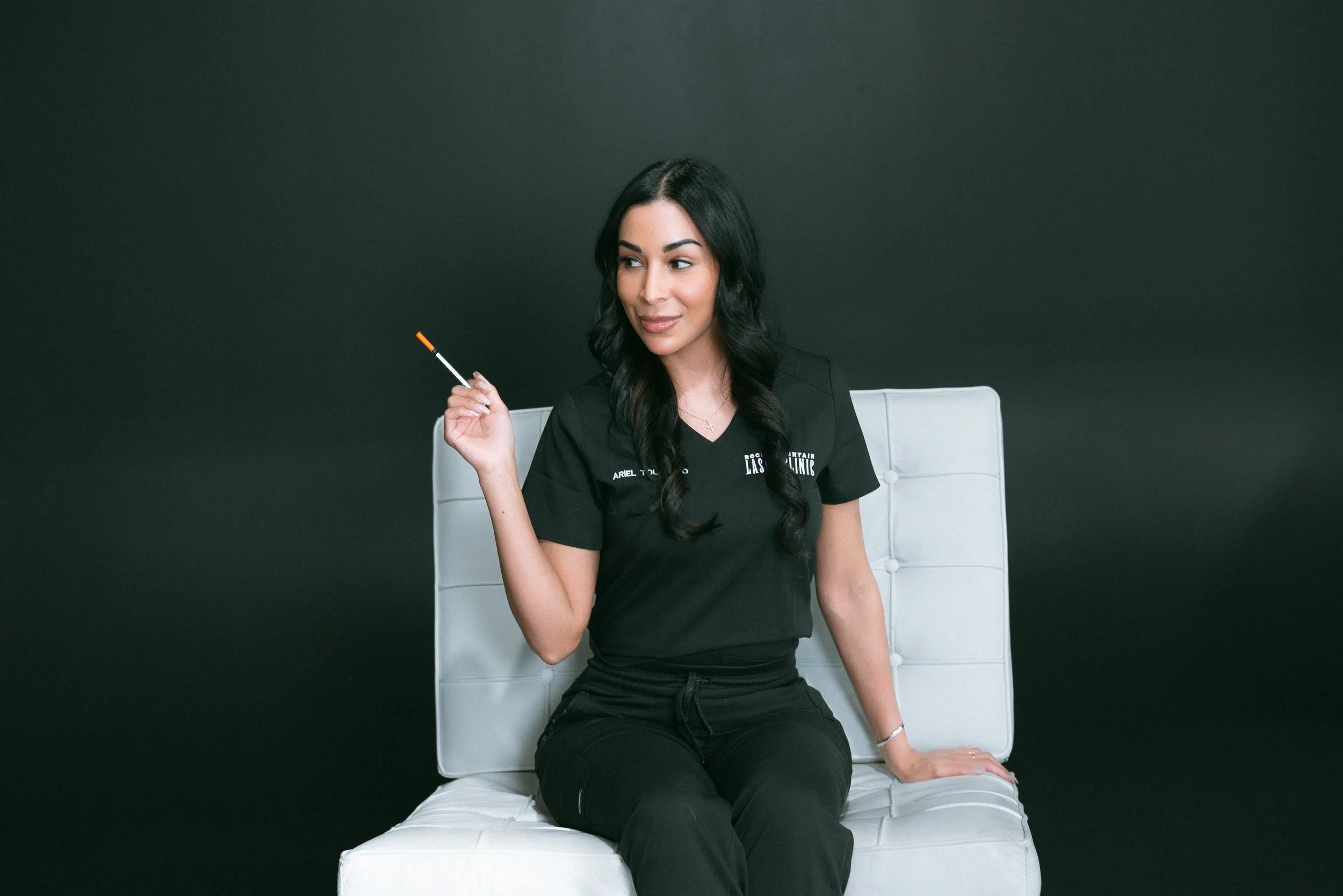 A woman with dark hair sitting on a white chair against a dark background, holding a cigarette in her right hand, wearing a black uniform with a name tag and logo, smiling softly.