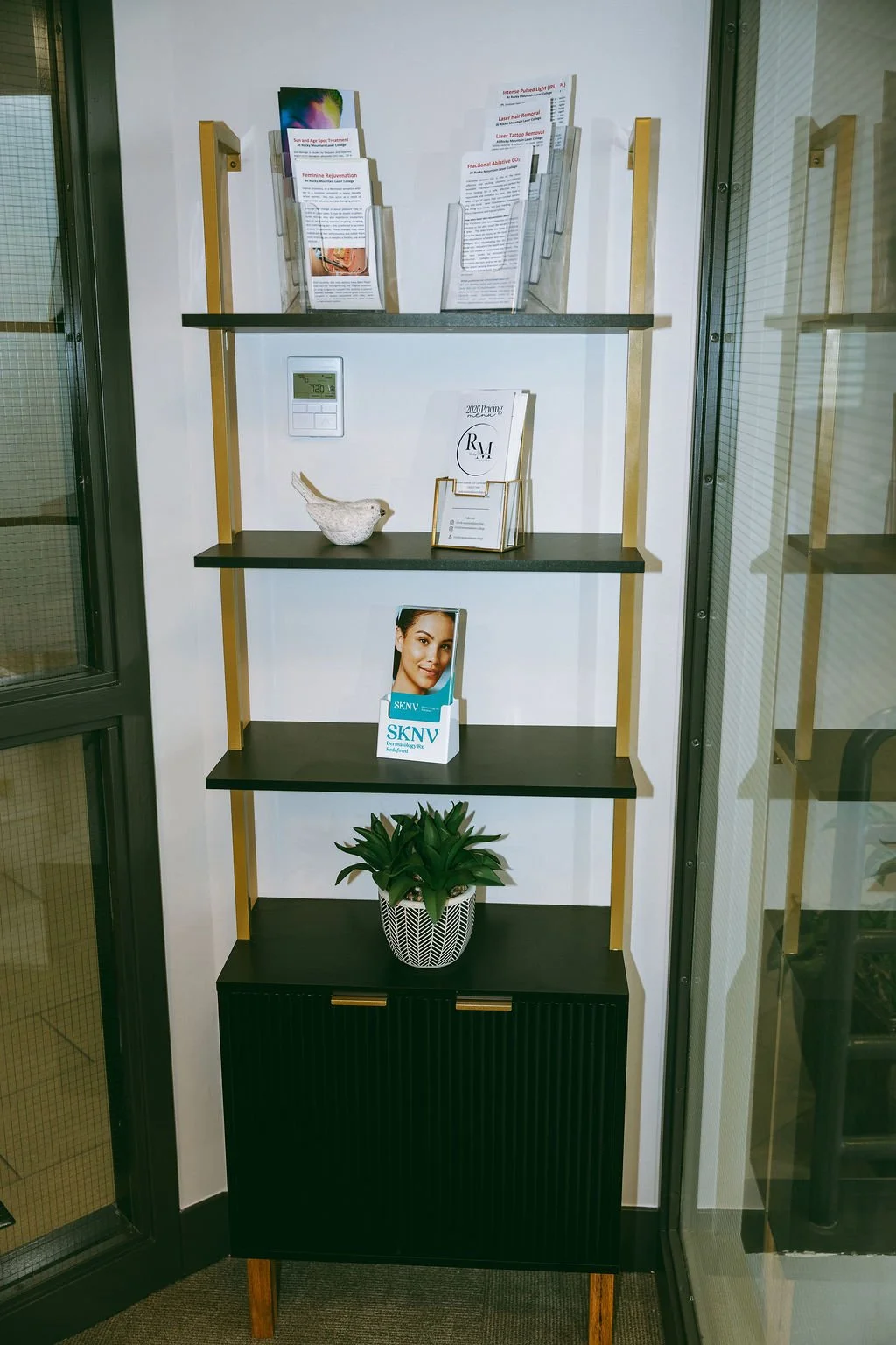 A modern black and gold shelving unit with four shelves, holding brochures, a decorative bowl, a pamphlet holder, a skincare product display, and a potted plant, situated next to a glass door.