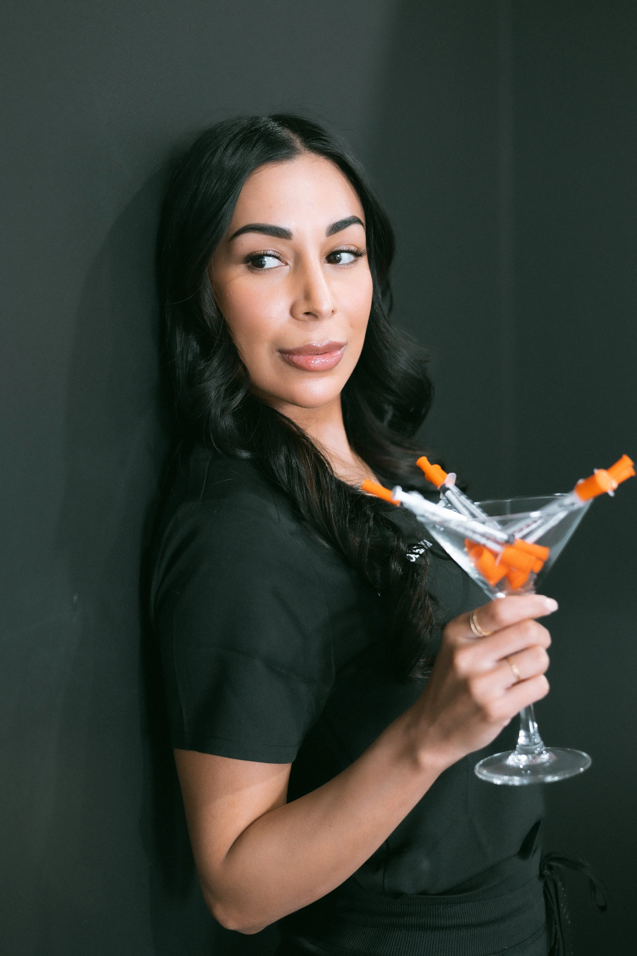 A woman with long black hair and peach-toned makeup is holding a martini glass filled with syringes.
