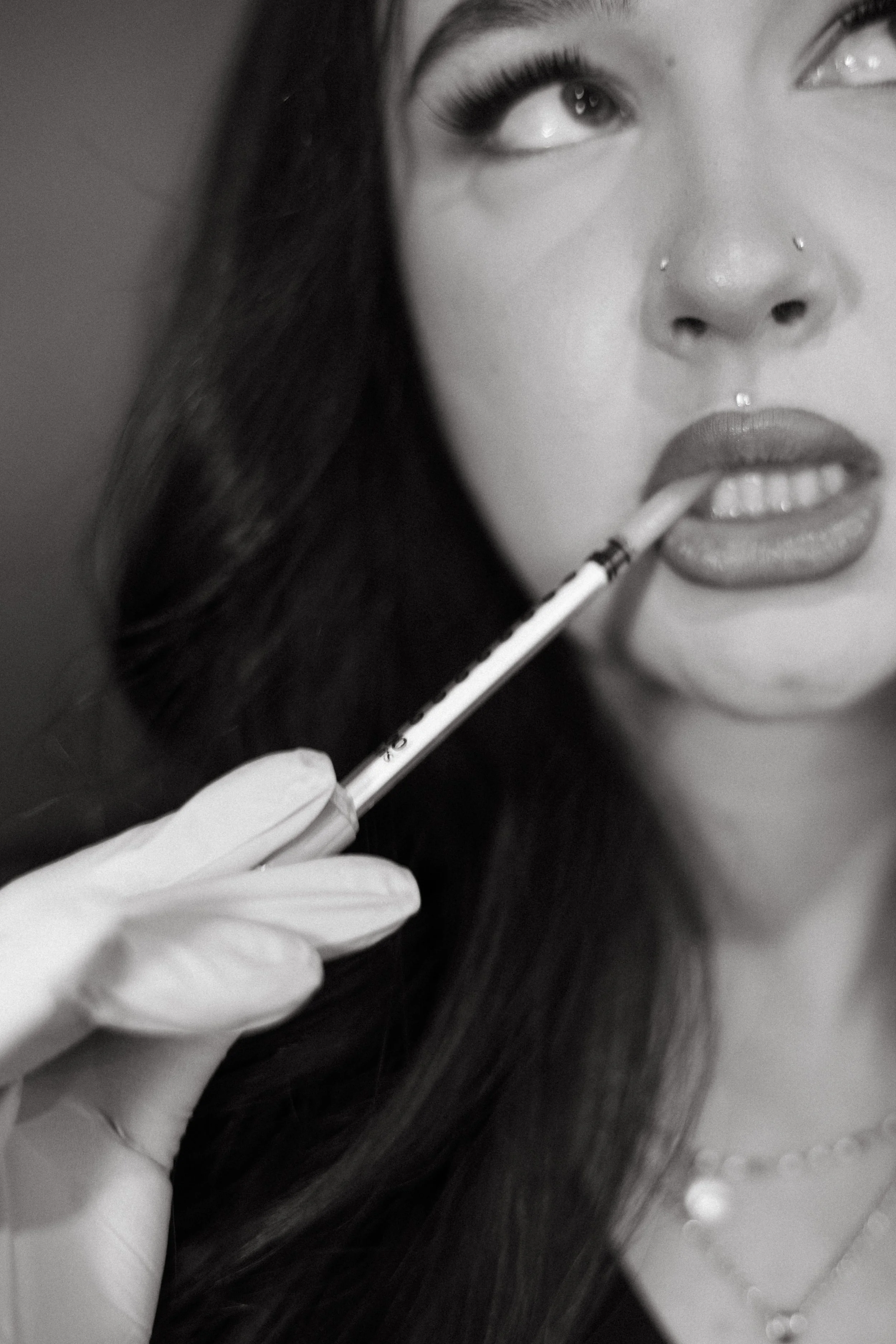 A woman with long dark hair holding a syringe near her lips, wearing gloves, and looking upward.