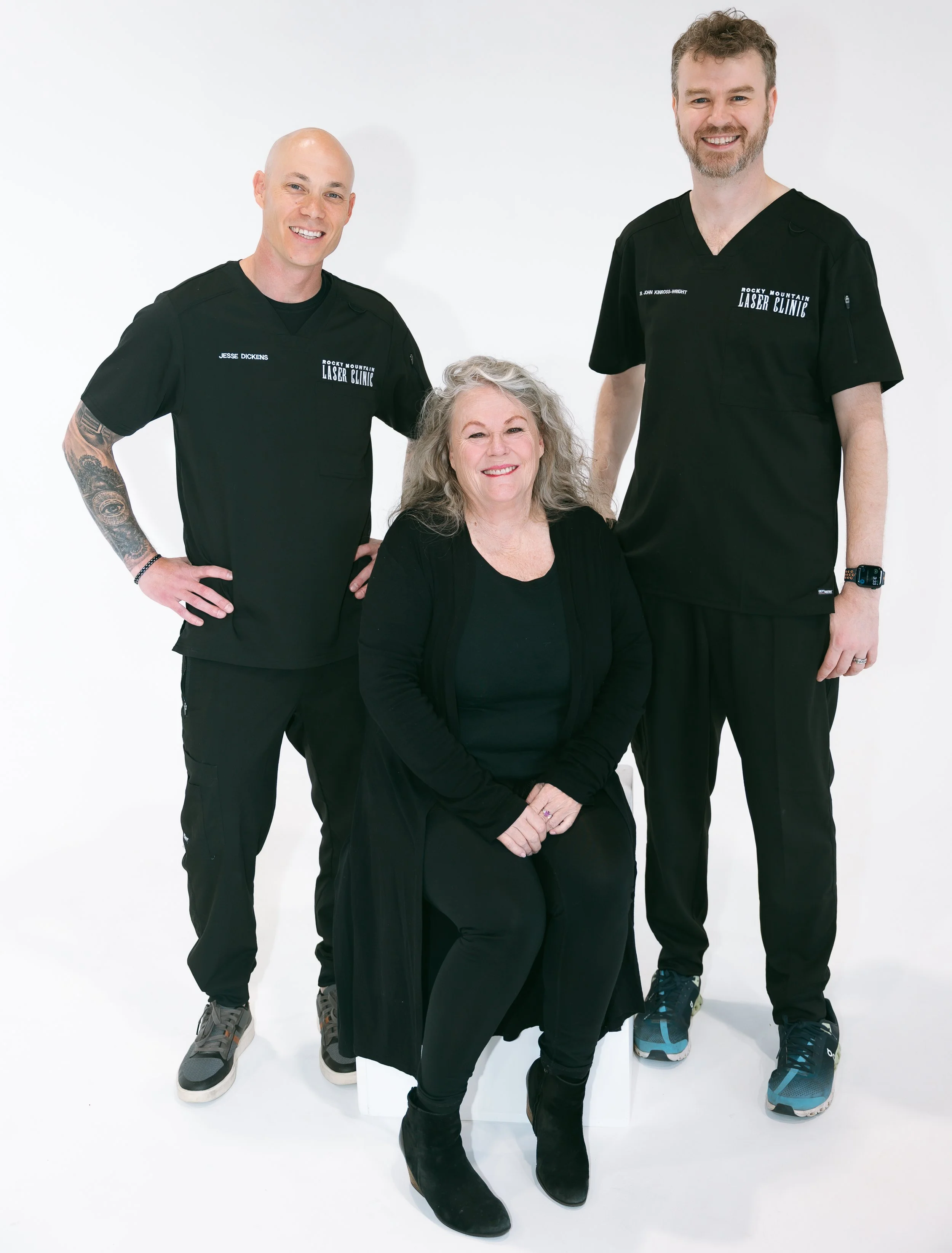 Three people, two men and one woman, standing and sitting against a white background. The woman is sitting in the center, smiling with gray curly hair, wearing a black top and black pants. The man on the left is bald with a tattoo on his right arm, wearing black medical scrubs with name tag "Jesse Dickens" and "Rocky Mountain Laser Clinic." The man on the right has short curly hair and a beard, wearing black medical scrubs with the name tag "John Kinnes-Wright" and a smartwatch on his left wrist. All three are smiling.