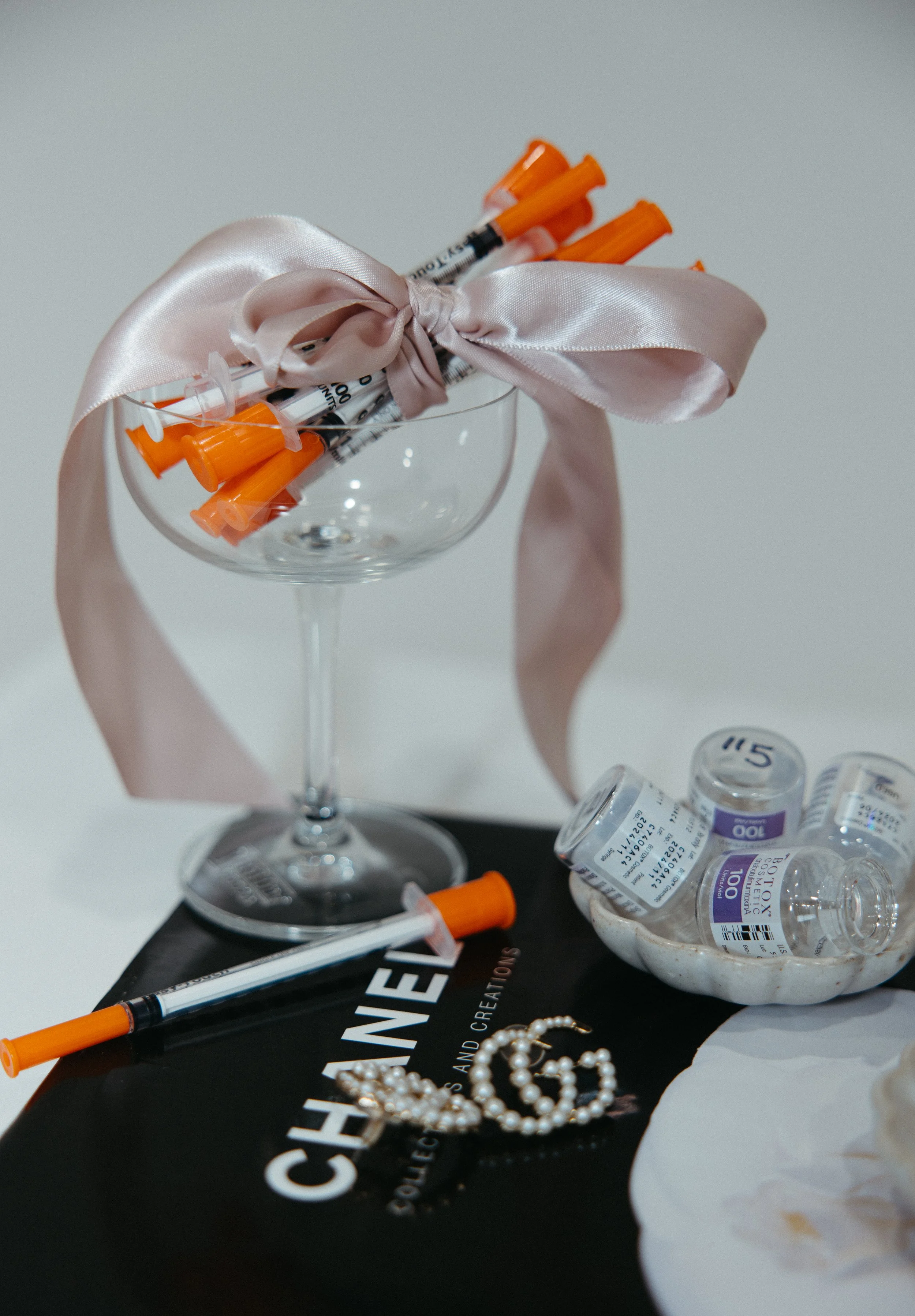 A champagne glass with a pink satin ribbon tied around its rim, holding several syringes with orange caps inside, with scattered prescription pill bottles, a black box, and pearl jewelry on a white surface.