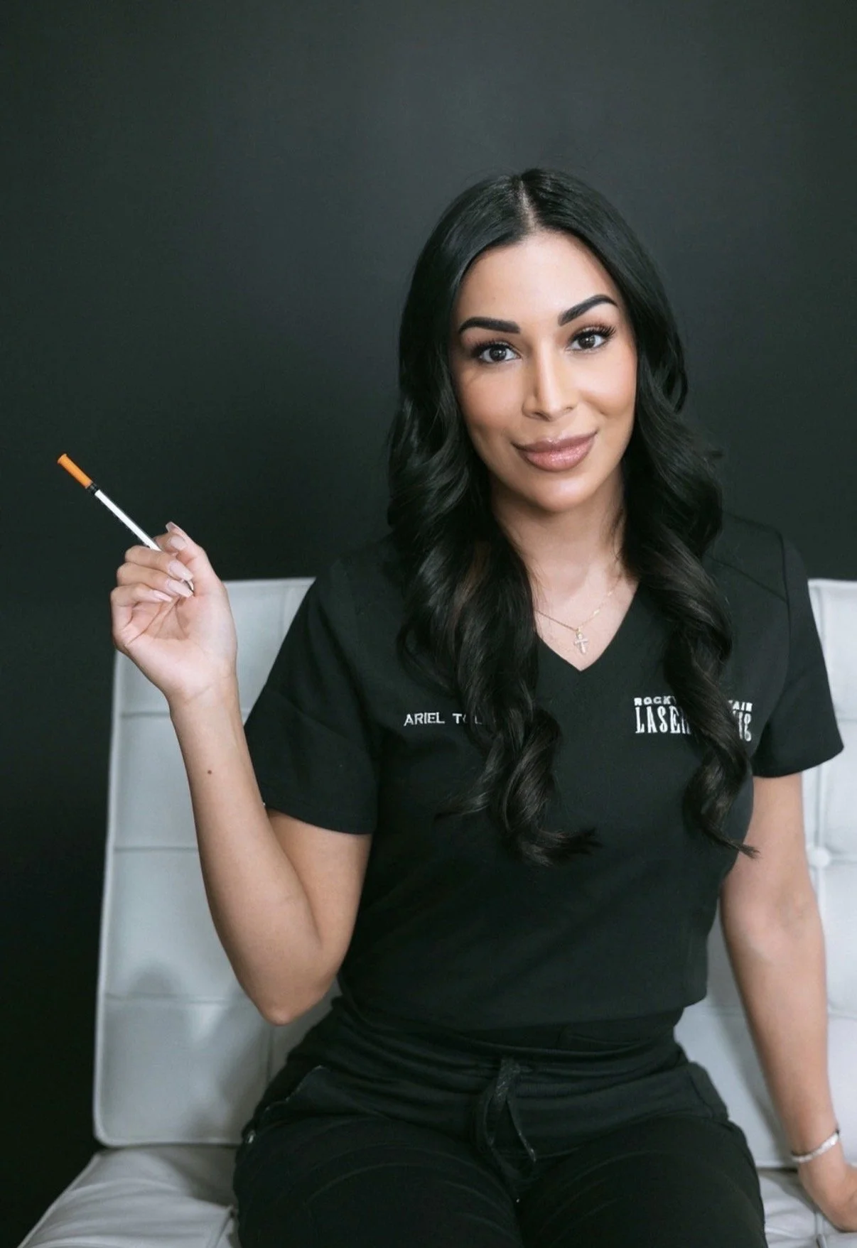 A woman with long black hair sitting on a white chair against a dark background, wearing a black medical or dental uniform, holding a syringe in her right hand, and smiling at the camera.