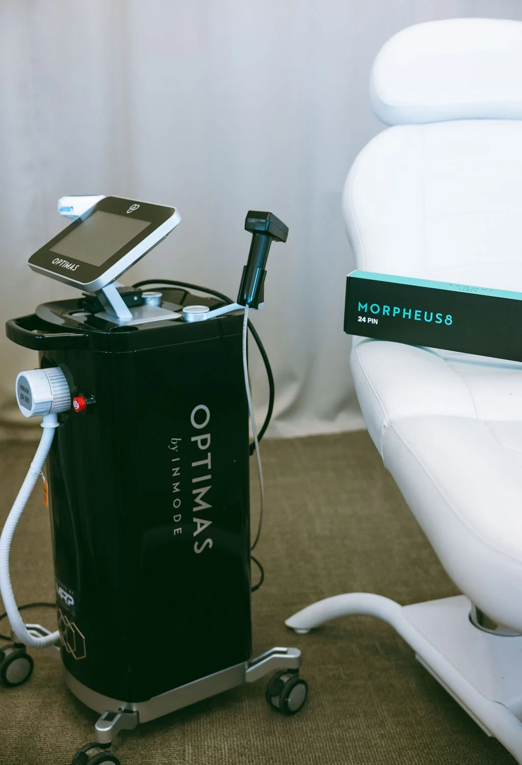 Medical equipment and a treatment chair in a clinic. The equipment has the word 'Optimas by Inmode' on it, and there is a box labeled 'Morpheus 8 24 PIN' on the chair.