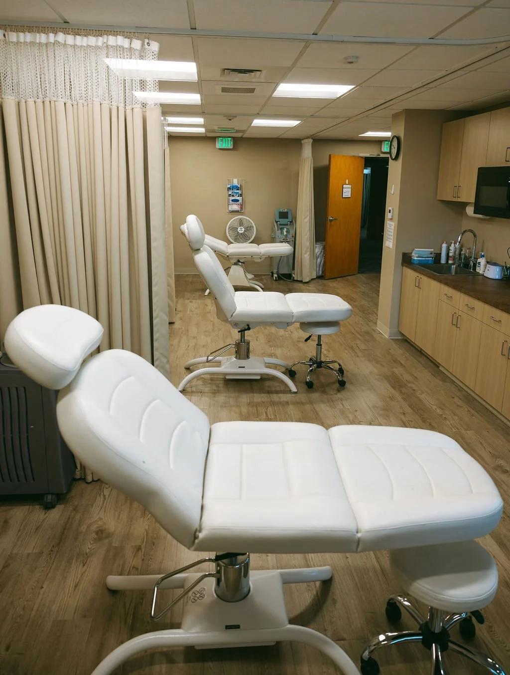Empty medical examination room with white adjustable examination chairs, a privacy curtain, medical equipment, and wooden cabinets.