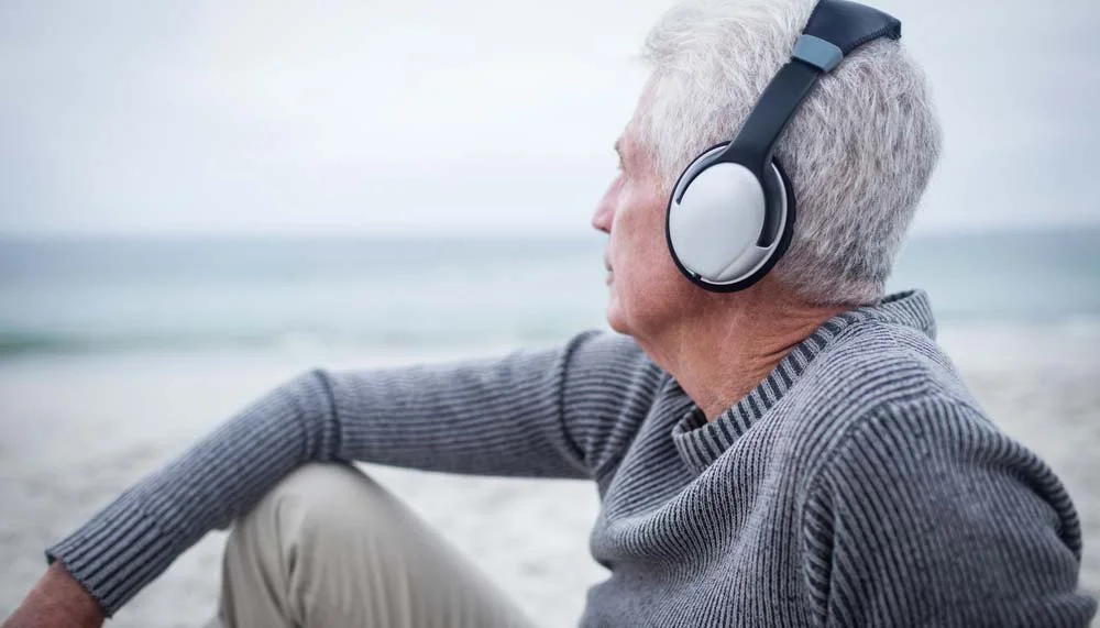 An elderly man with gray hair sitting on a beach, wearing headphones and a gray sweater, looking out at the ocean.