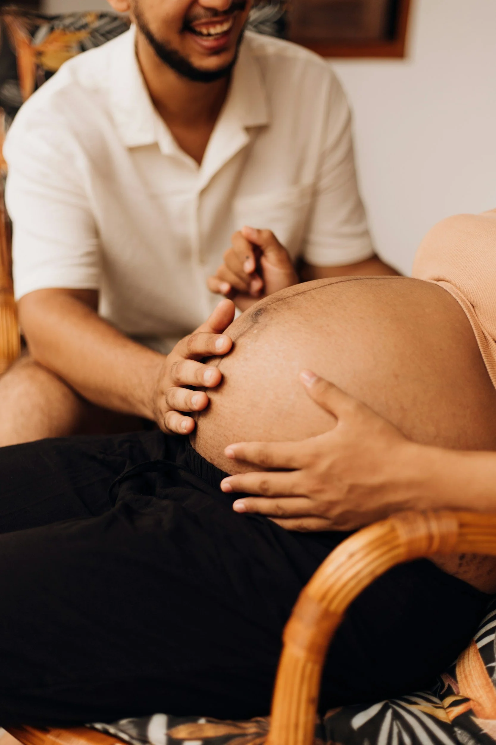 A man smiling and holding a pregnant woman's belly as she lies on a chair.