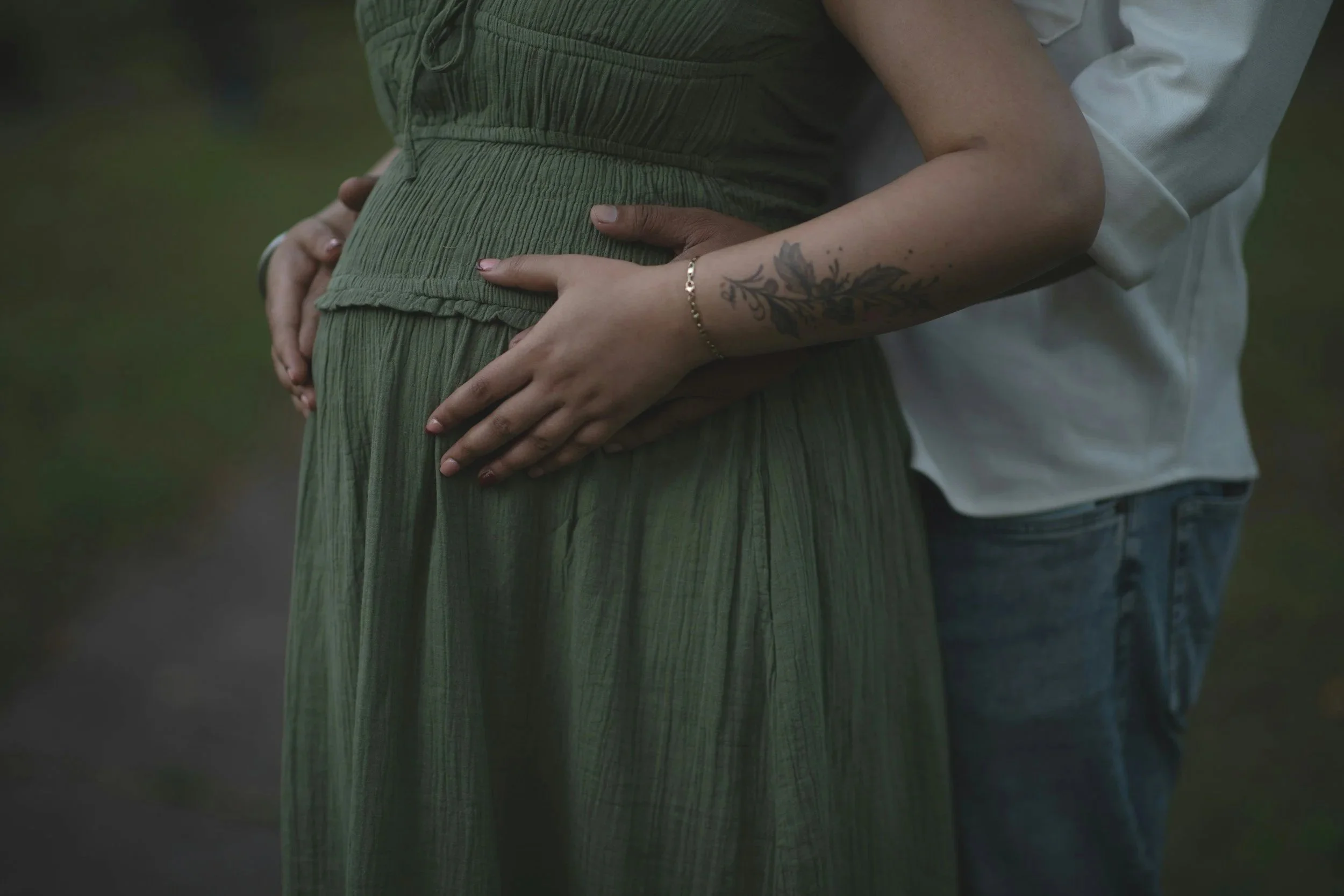 A person wearing a green dress is holding their pregnant belly with their hands. Another person, in a white shirt, is standing behind them with their arms around the pregnant person. The second person's hand is also on the belly. The background is bl