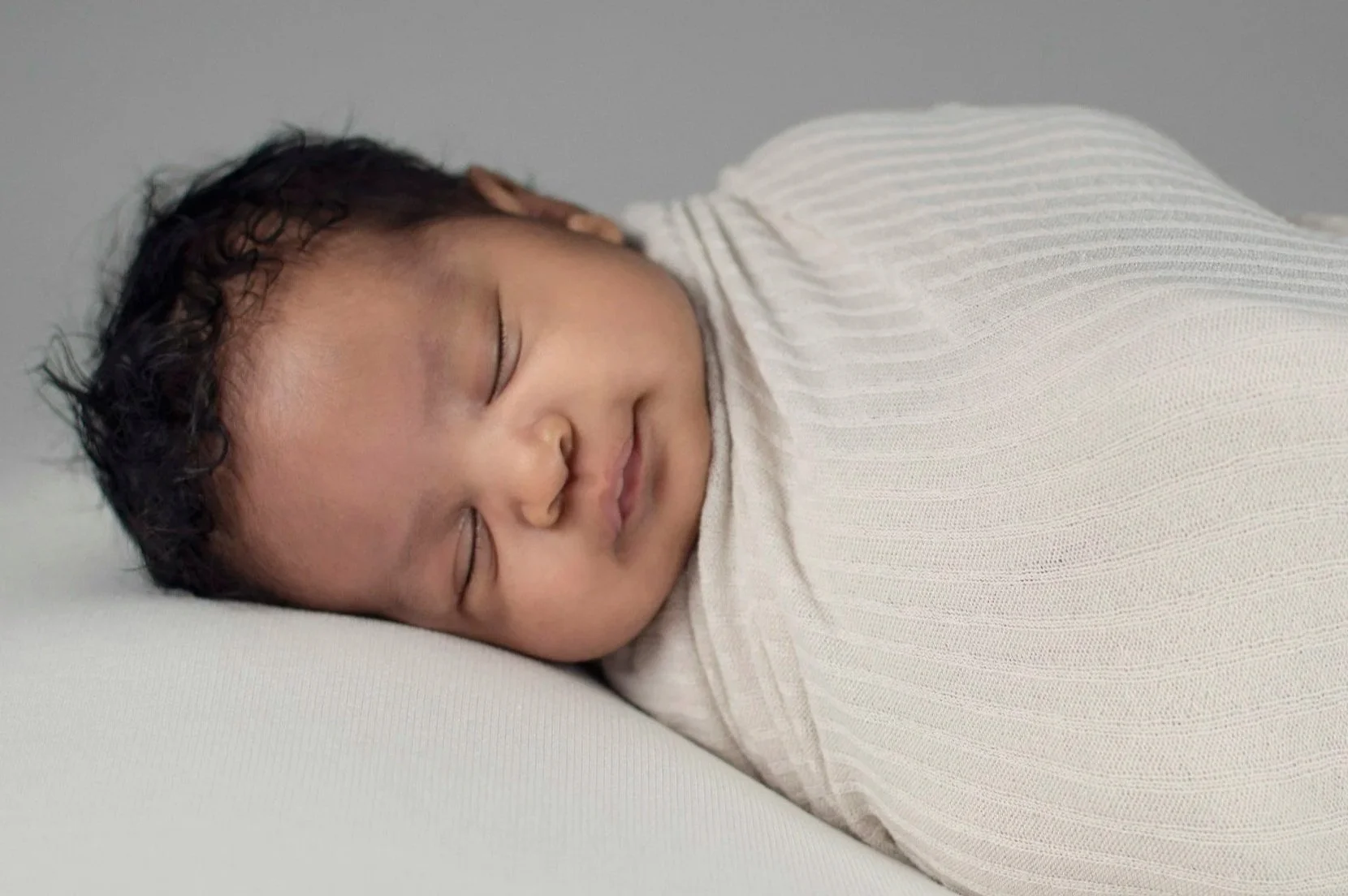 A sleeping baby with curly black hair, resting on a white pillow, wearing a off-white, ribbed long-sleeve shirt.