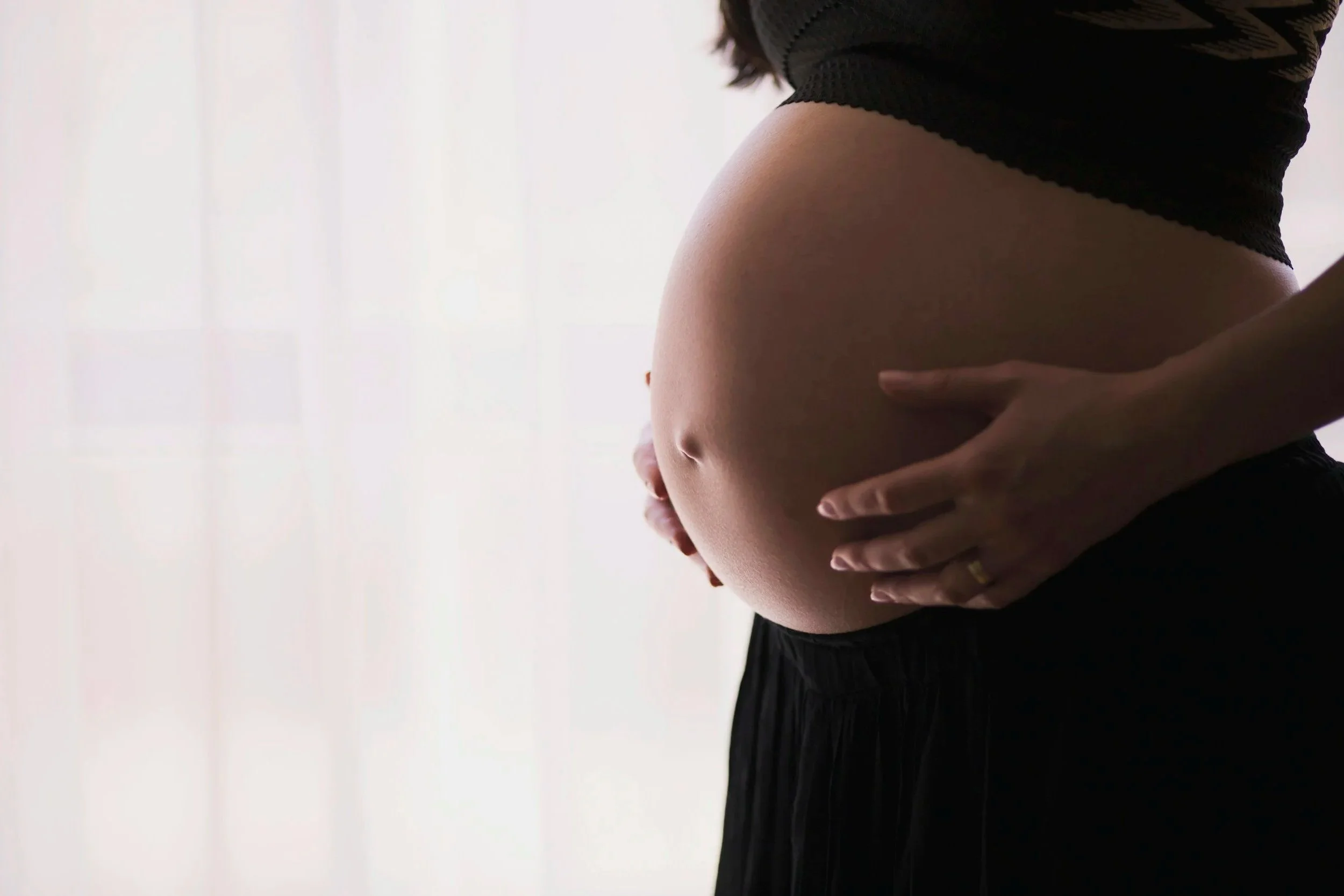 Close-up of a pregnant woman holding her belly, wearing a black top and black skirt, with her hands gently resting on her abdomen.