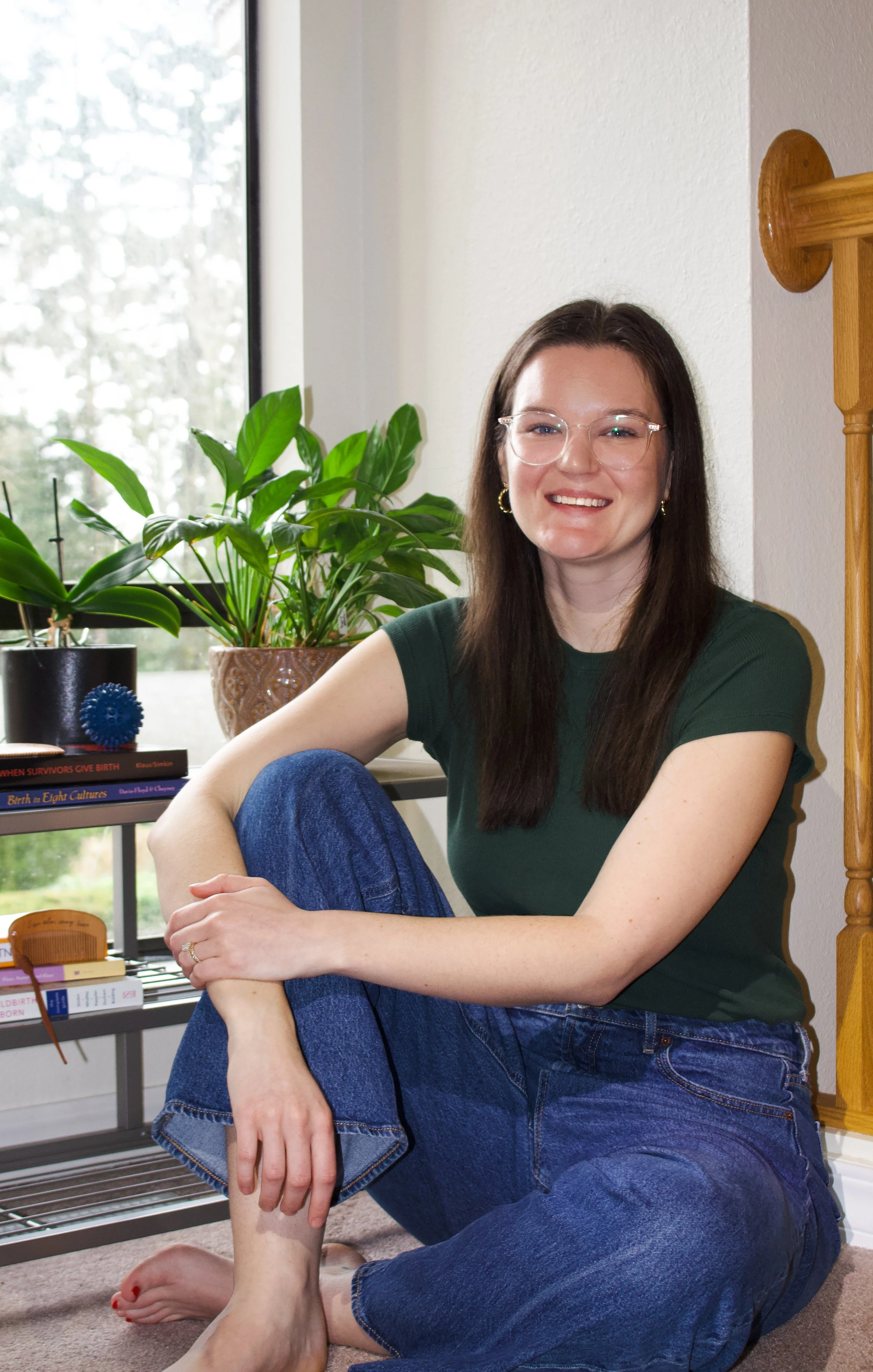 A young woman with glass glasses and long brown hair sitting on the floor, smiling, next to a window with green plants and a bookshelf.