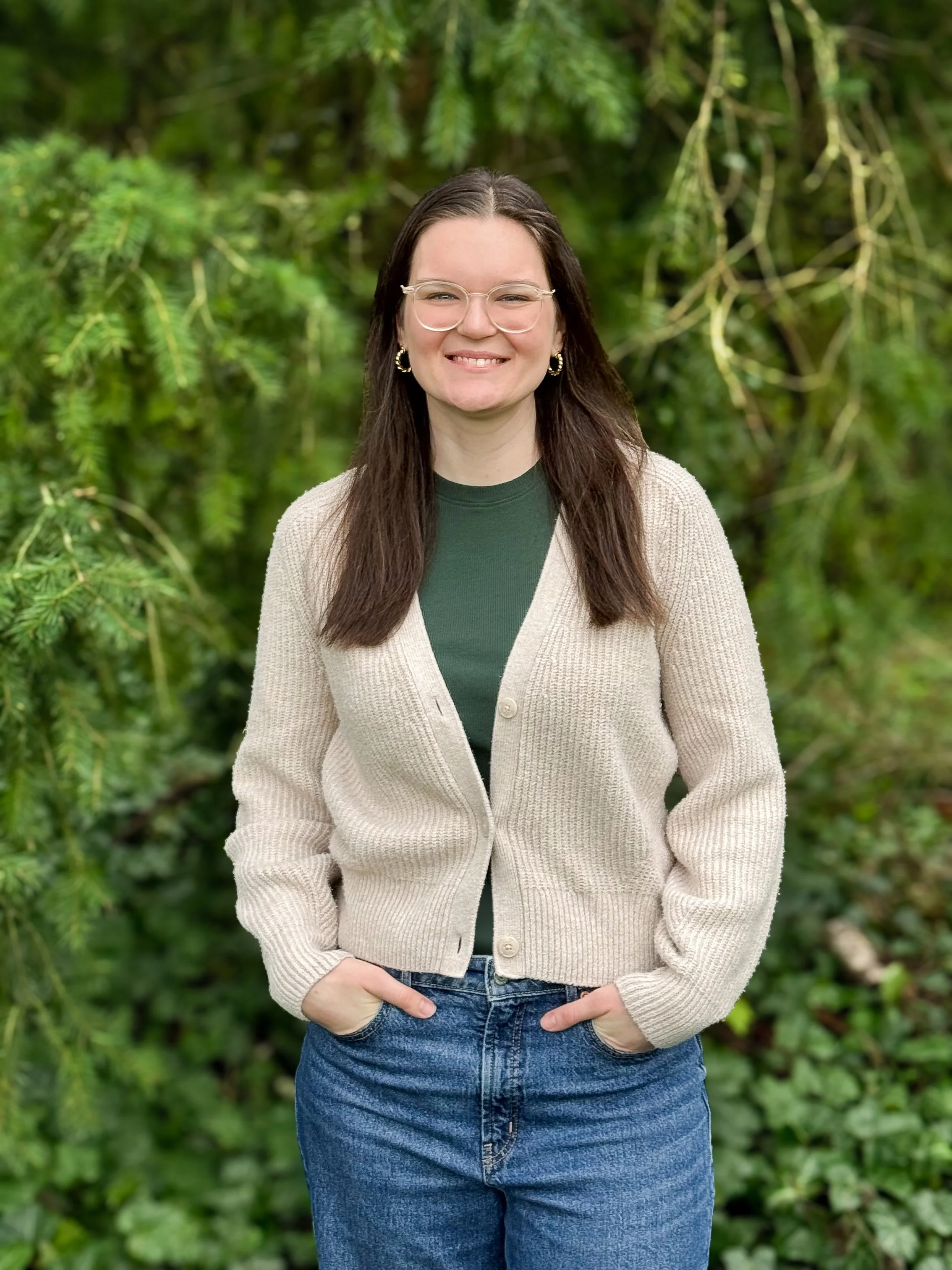 Smiling woman with long dark hair, glasses, gold hoop earrings, wearing a beige cardigan, green top, and blue jeans, standing outdoors with green foliage background.