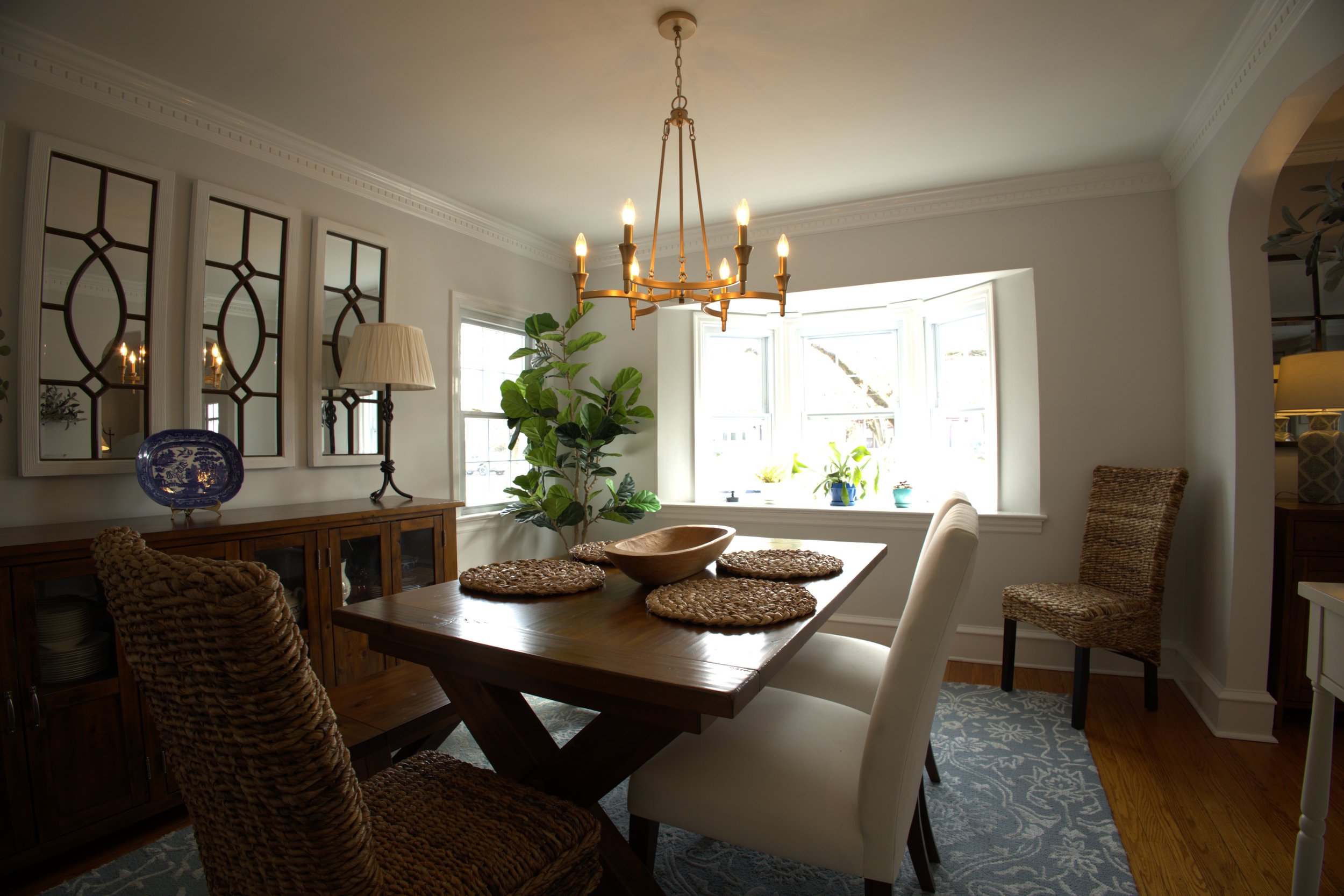 A cozy dining room with a wooden table, four chairs, a chandelier, a sideboard, a large window with plants on the sill, and decorative wall mirrors.