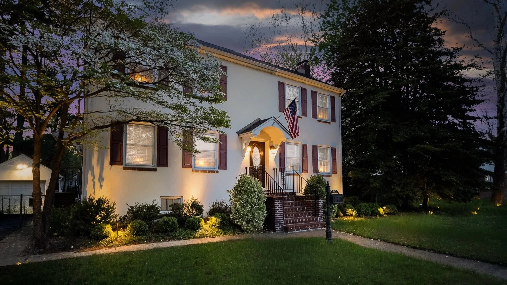 A two-story white house with red shutters, a front porch, and an American flag, illuminated at dusk with trees and a garden in front.
