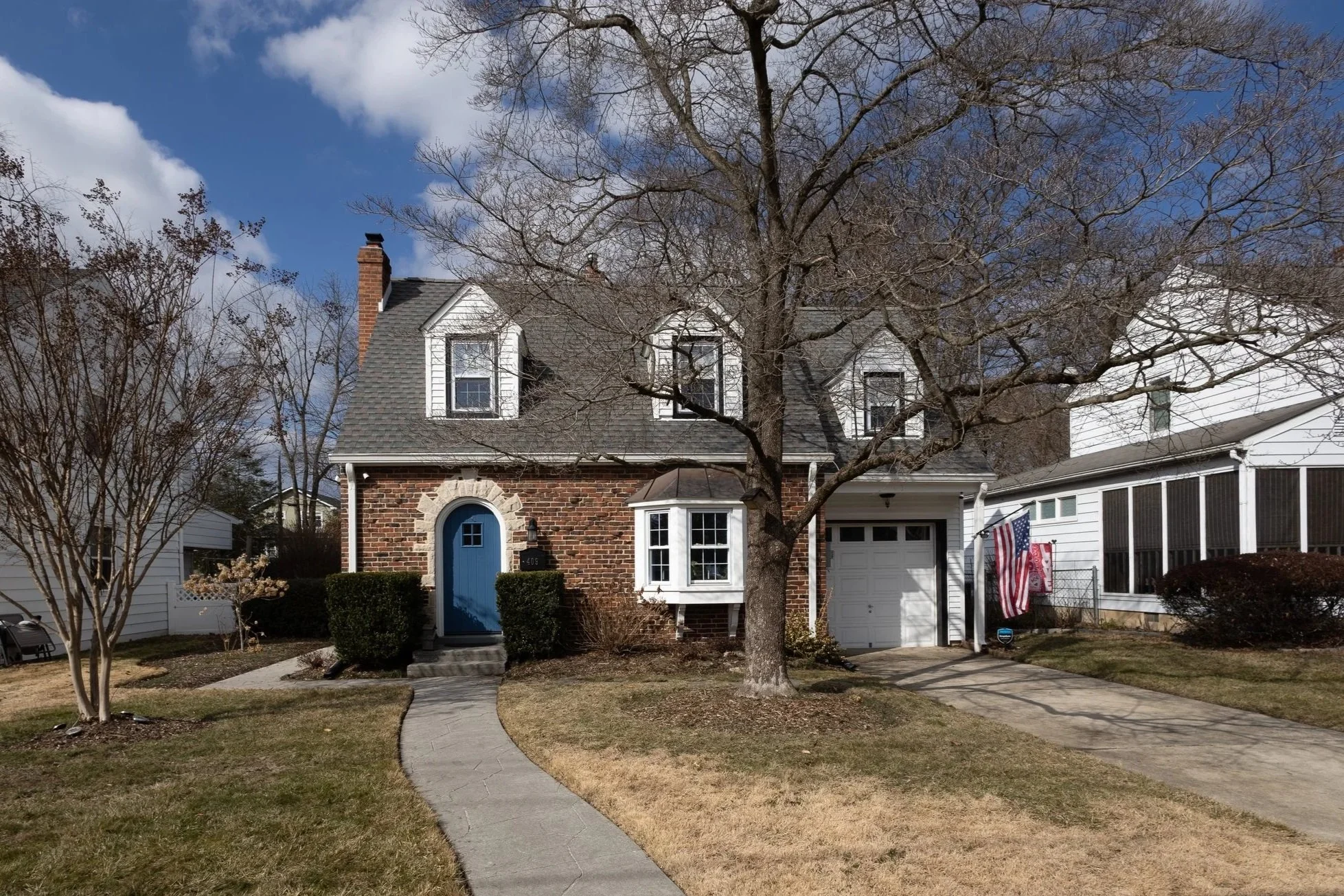 A two-story house with a brick facade, a blue front door, and a small front yard with grass and a tree. There is a sidewalk leading to the front door and a driveway next to the house. The house has three dormer windows and a chimney. The sky is partly cloudy.