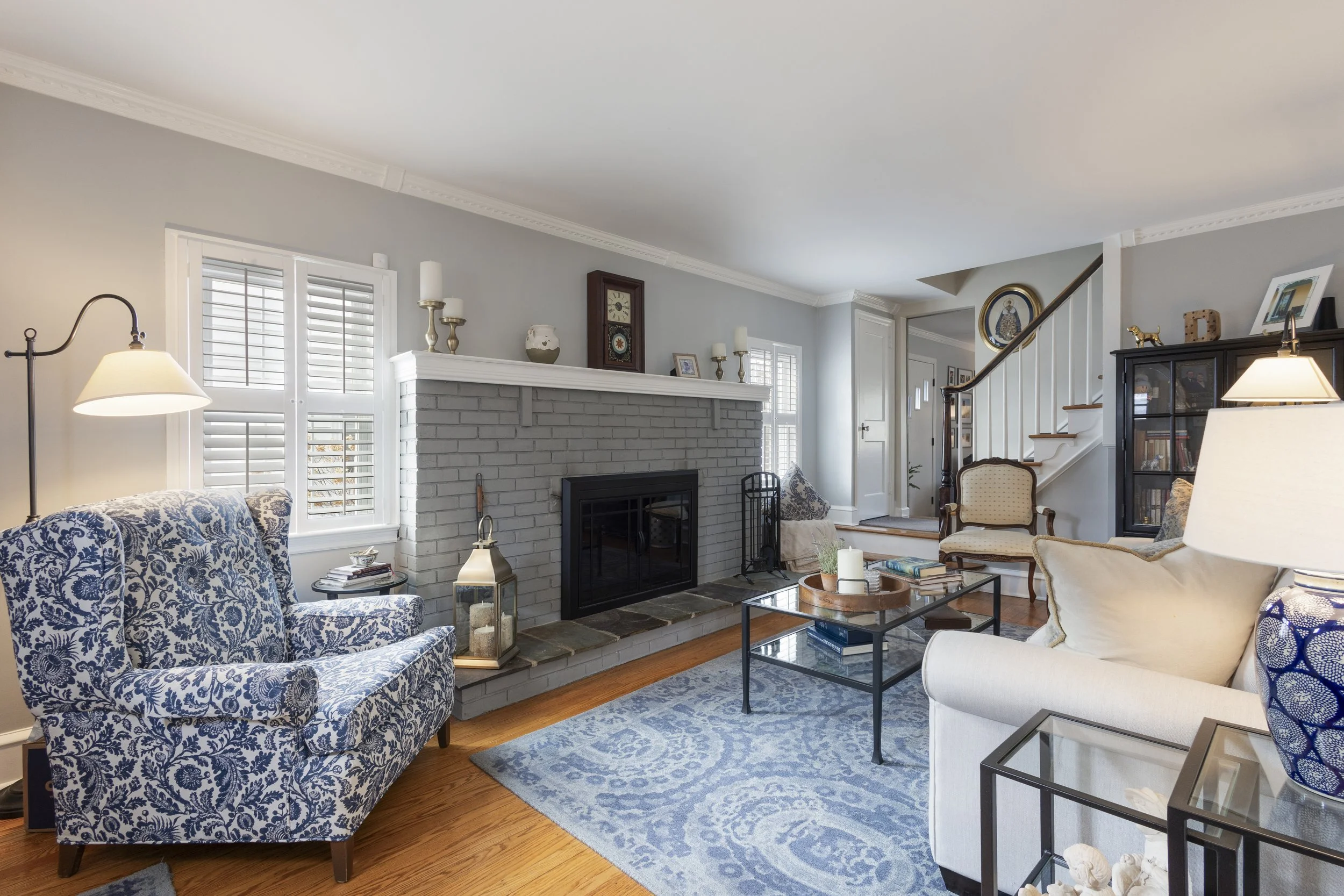 Living room with a gray brick fireplace, blue and white patterned armchair, white sofa, glass coffee table, and various decor items.