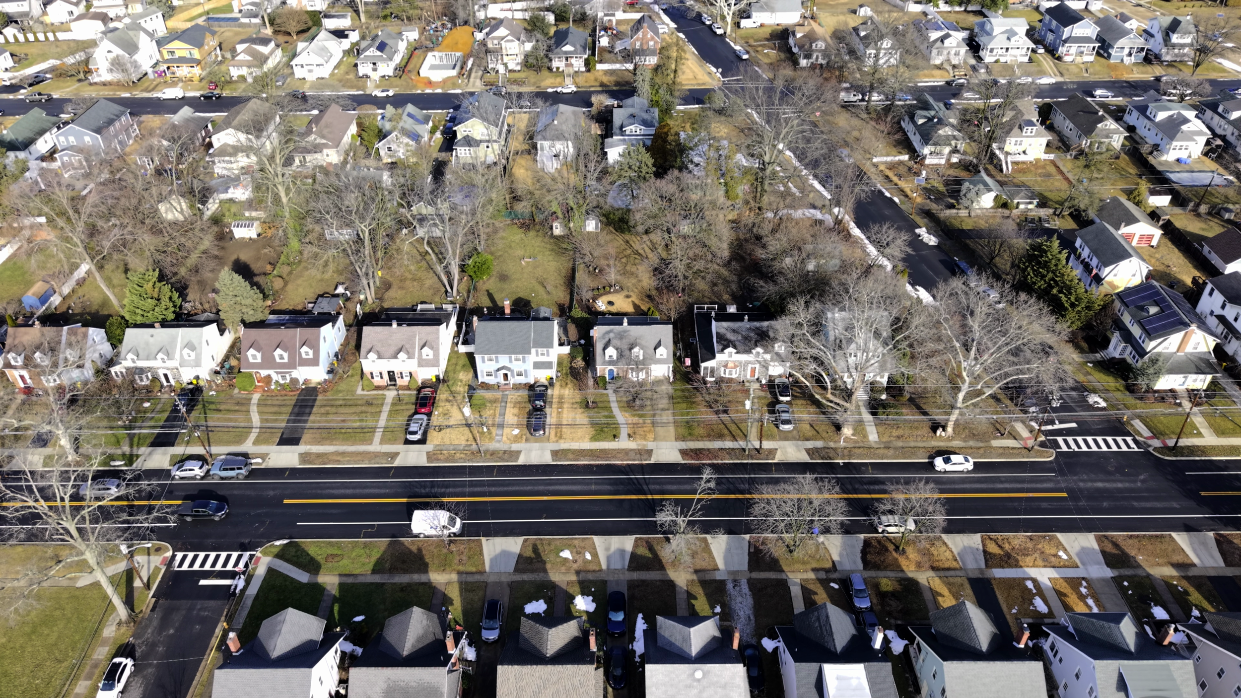 An aerial view of a residential neighborhood showing houses, trees, a street with cars, and sidewalks.