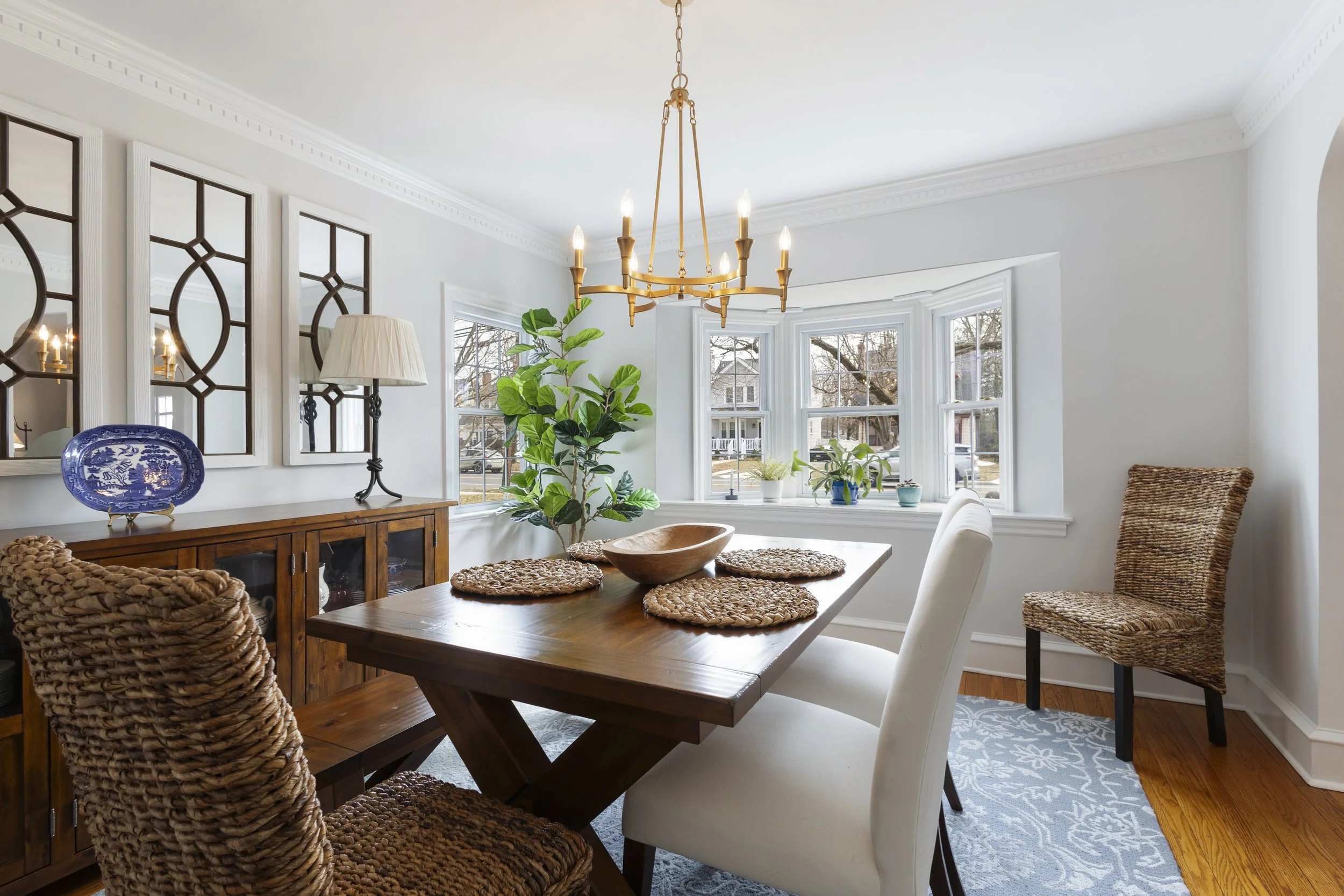 A bright dining room with a wooden dining table, six chairs, a chandelier, and window seats with potted plants, white walls, and hardwood floors.