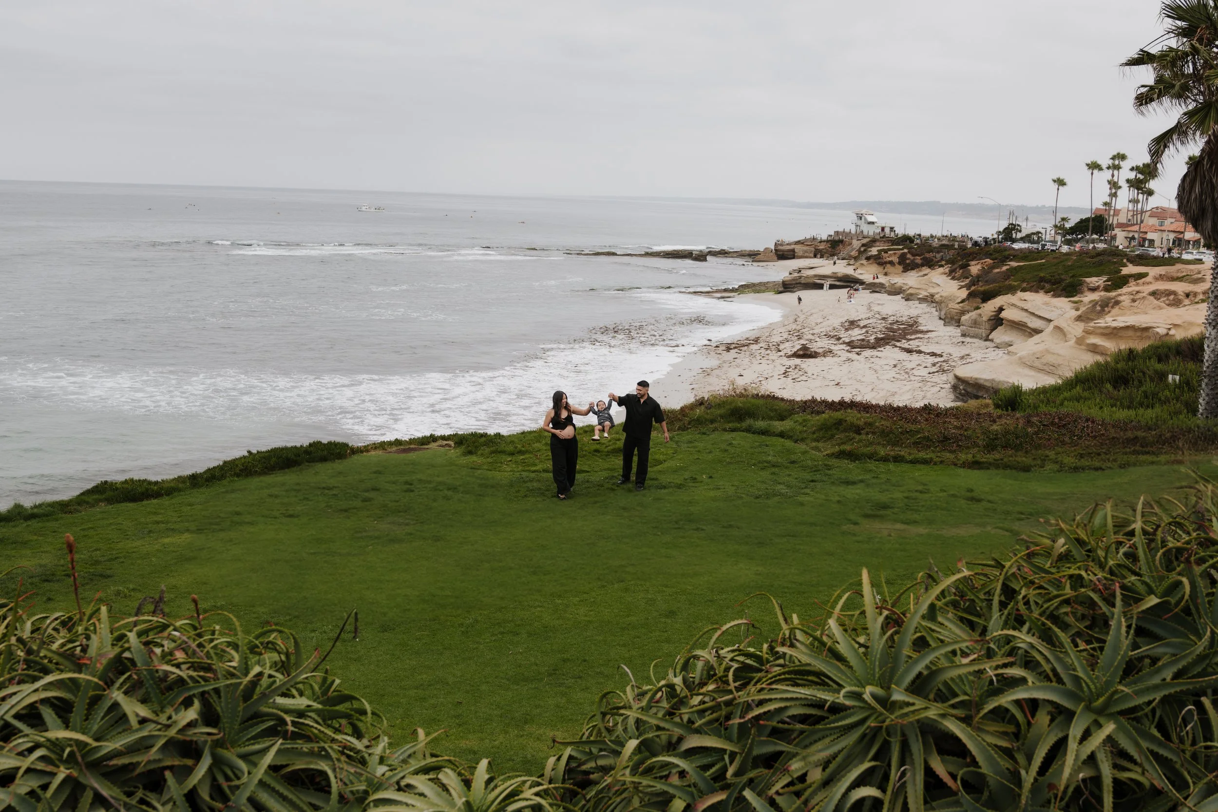 A family of three walking along a grassy area near the beach on a cloudy day, with the ocean, rocky shoreline, and palm trees in the background.