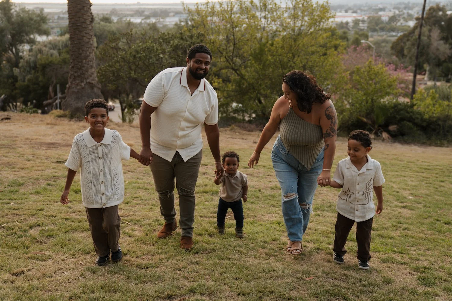 A family of six, two adults and four children, walks holding hands in a park-like setting with trees and grass, smiling and enjoying each other's company.