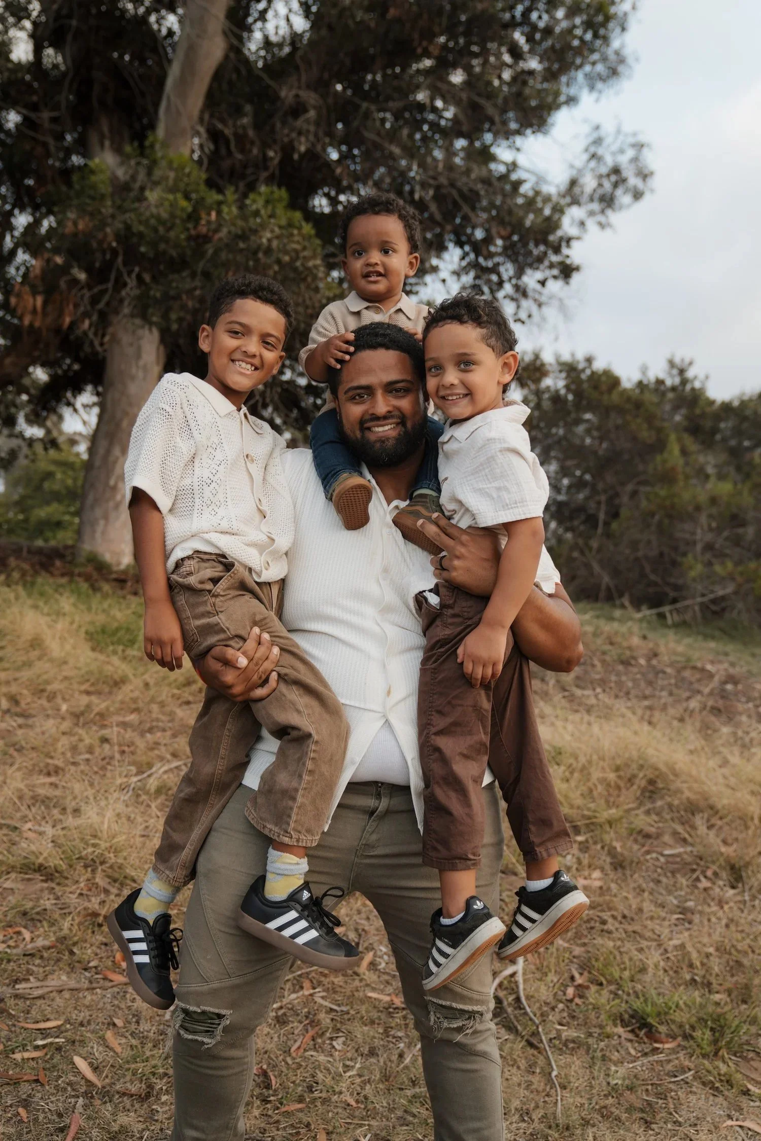 A man holding two young boys on his shoulders and standing in a park with trees in the background. All three are smiling and wearing casual clothes.