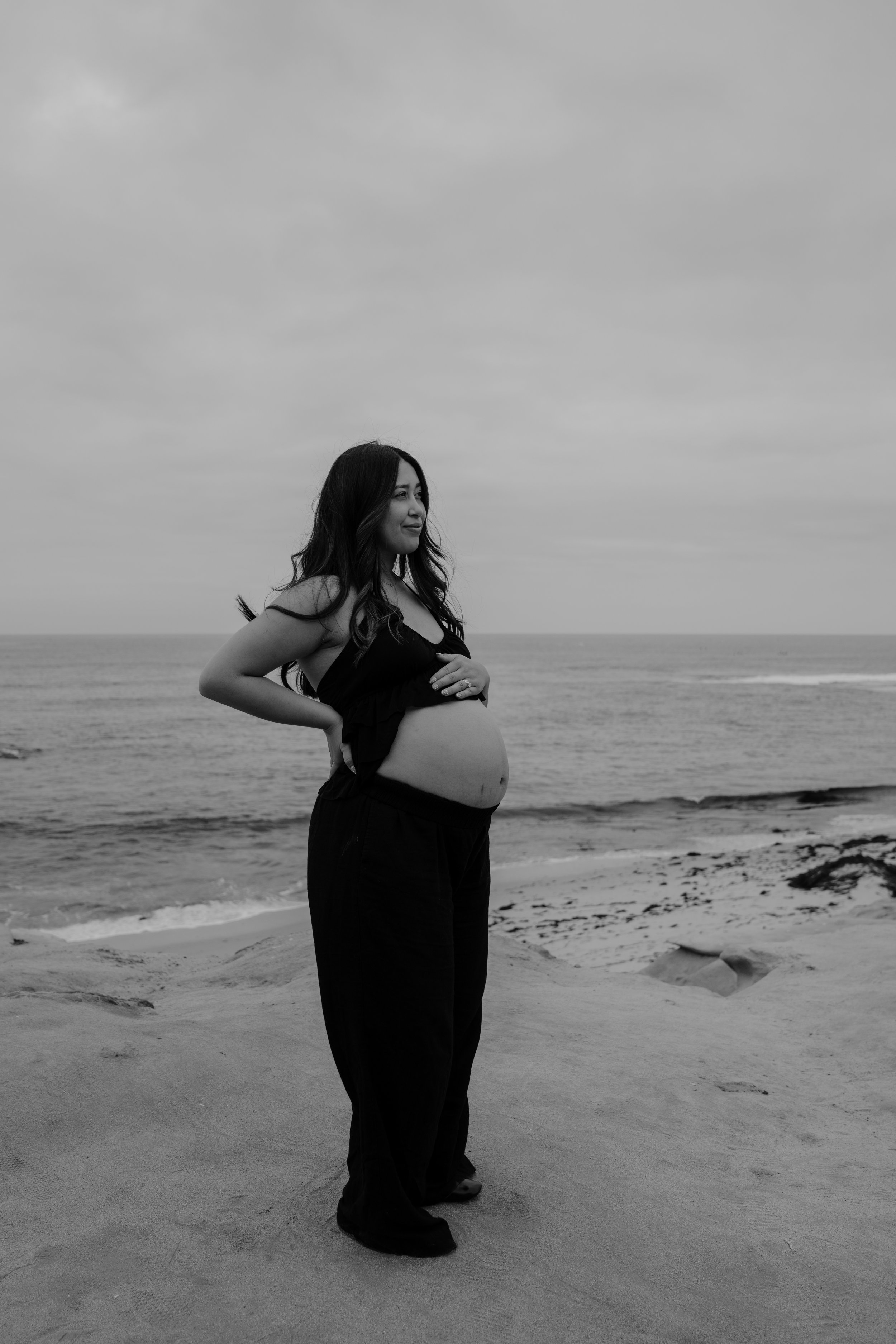 A pregnant woman stands on the beach, smiling, with her hands on her belly, looking out at the ocean under a cloudy sky.