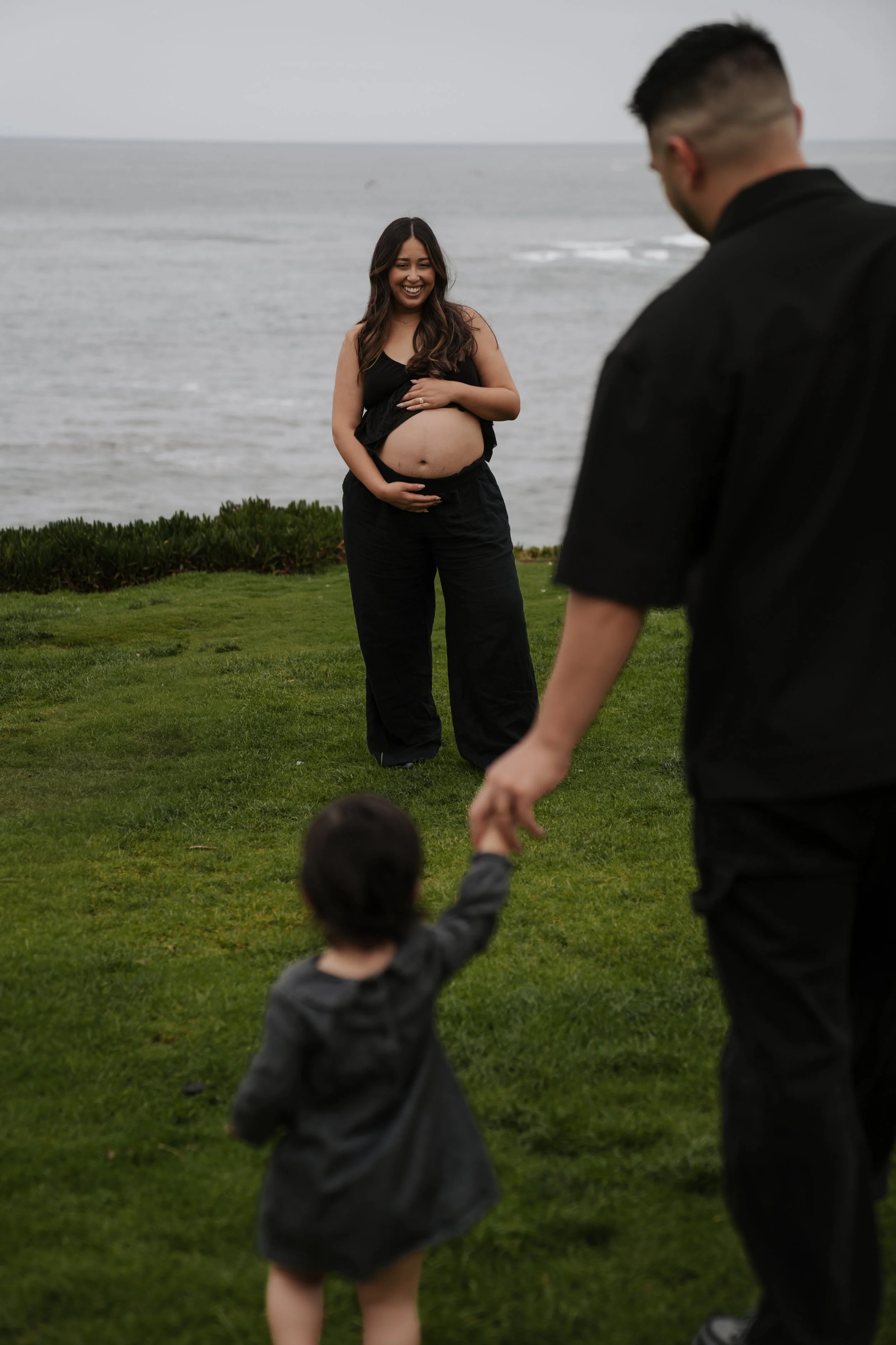 A pregnant woman smiling at a man and a child while standing on a grassy area near the ocean during daytime.