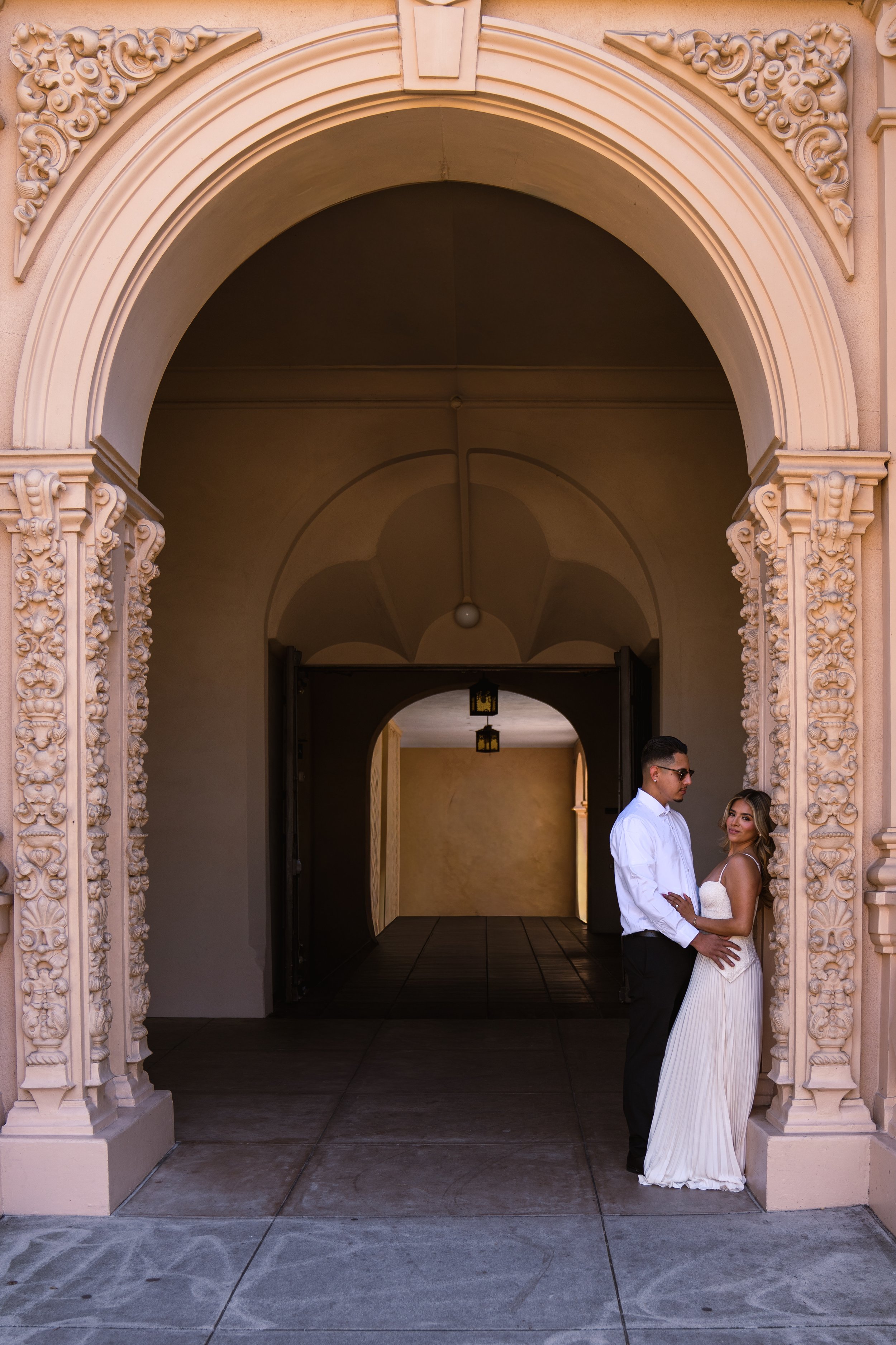 A bride and groom holding hands and standing under an ornate archway with intricate carvings on the sides and ceiling lights hanging in the background.